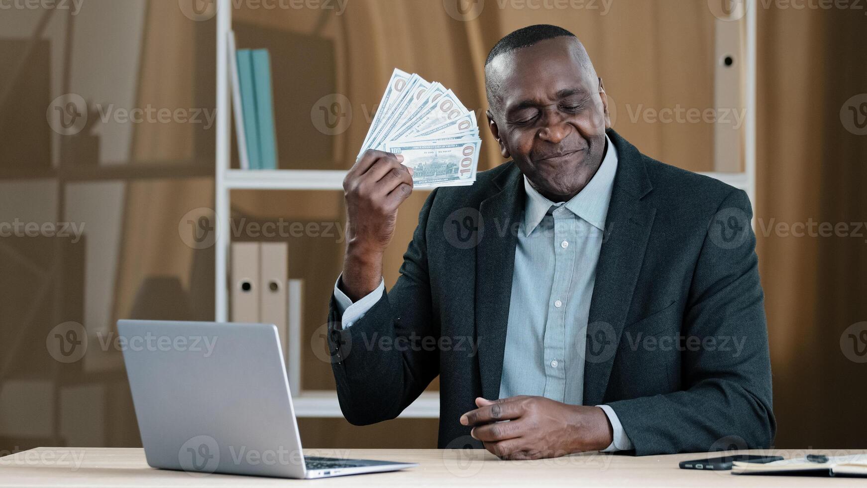 Old mature African businessman 60s worker banker banking agent senior man sitting in office rejoices in financial win holds fan of money sniffing dollars satisfied with salary loan wealth success photo