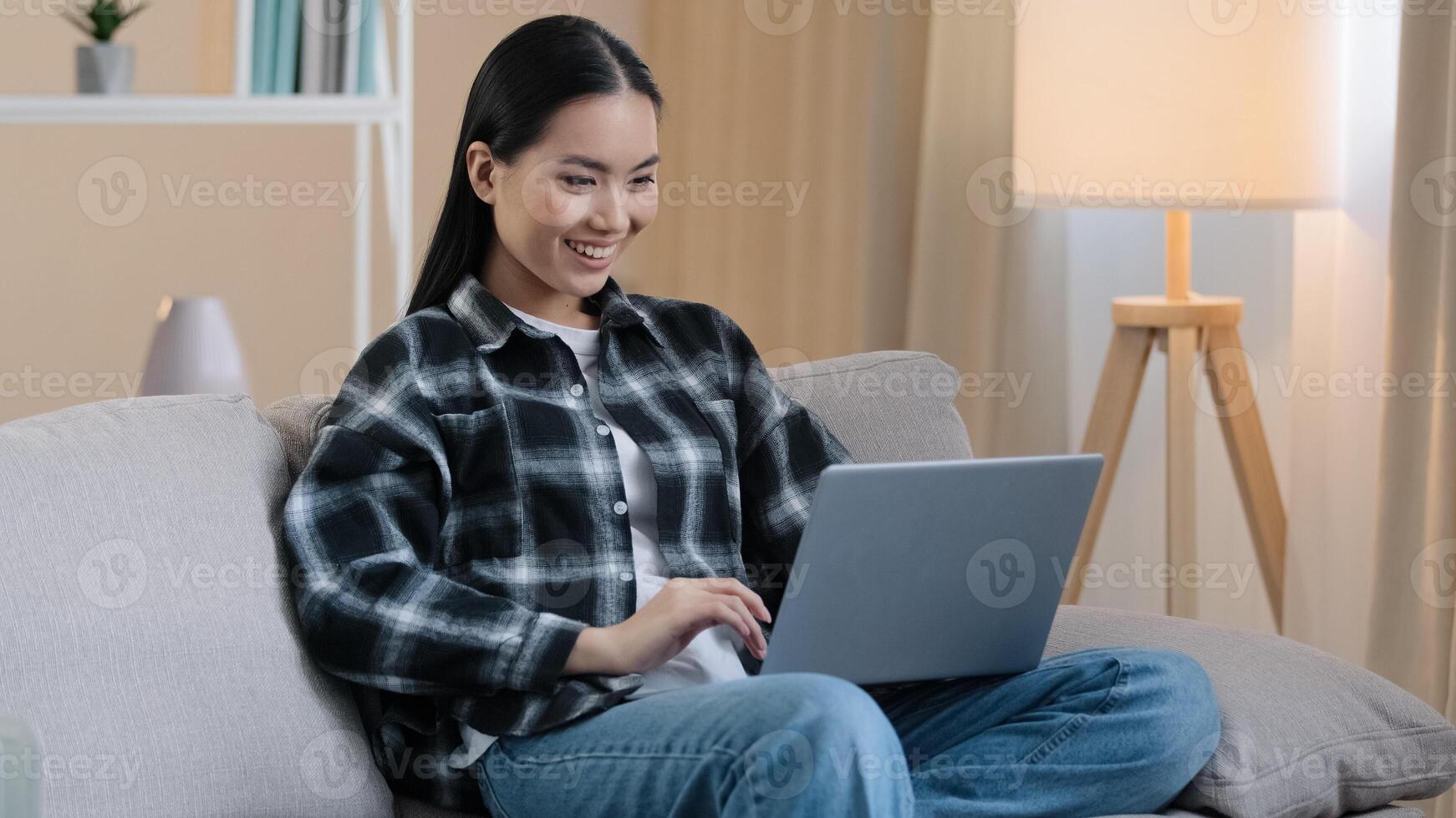 Asian 20s girl user millennial female student freelancer woman chatting internet with laptop home sofa playing computer game smiling happy choosing shopping watching funny social networks app photo
