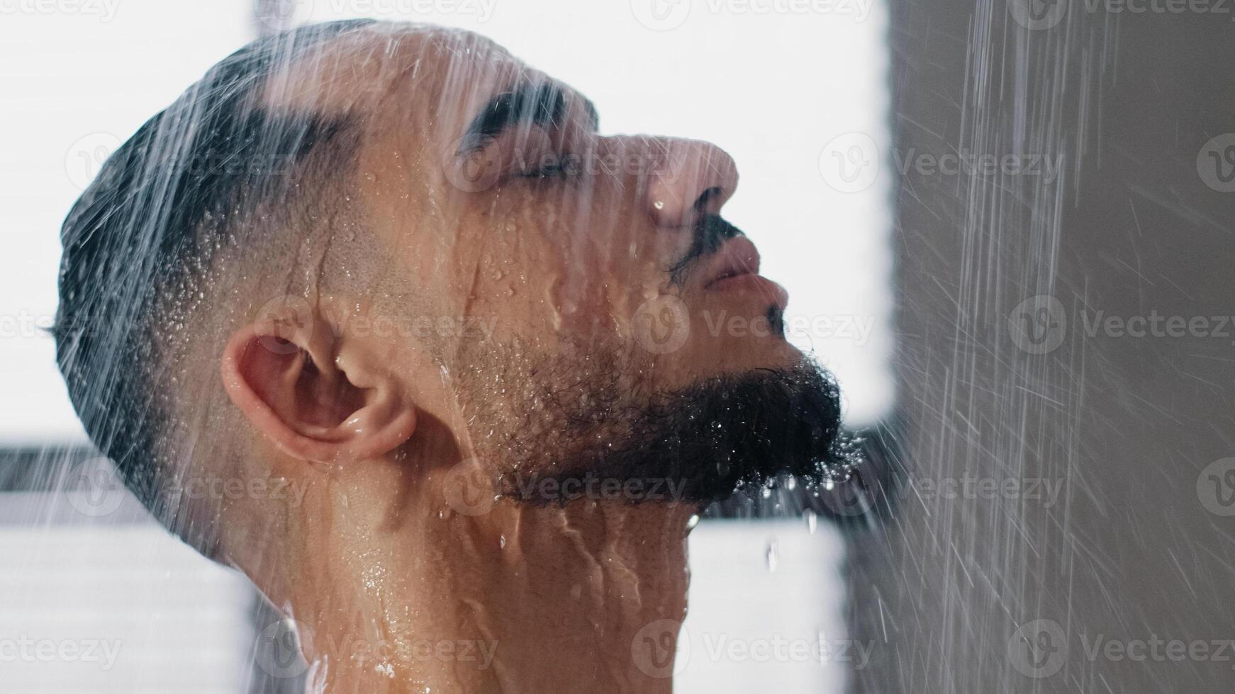 Sexy seductive arab bearded man in shower washing head showering in bathroom at home close up. Millennial brunette guy rinsing shampoo and conditioner from hair in warm bath hot water runs down face photo