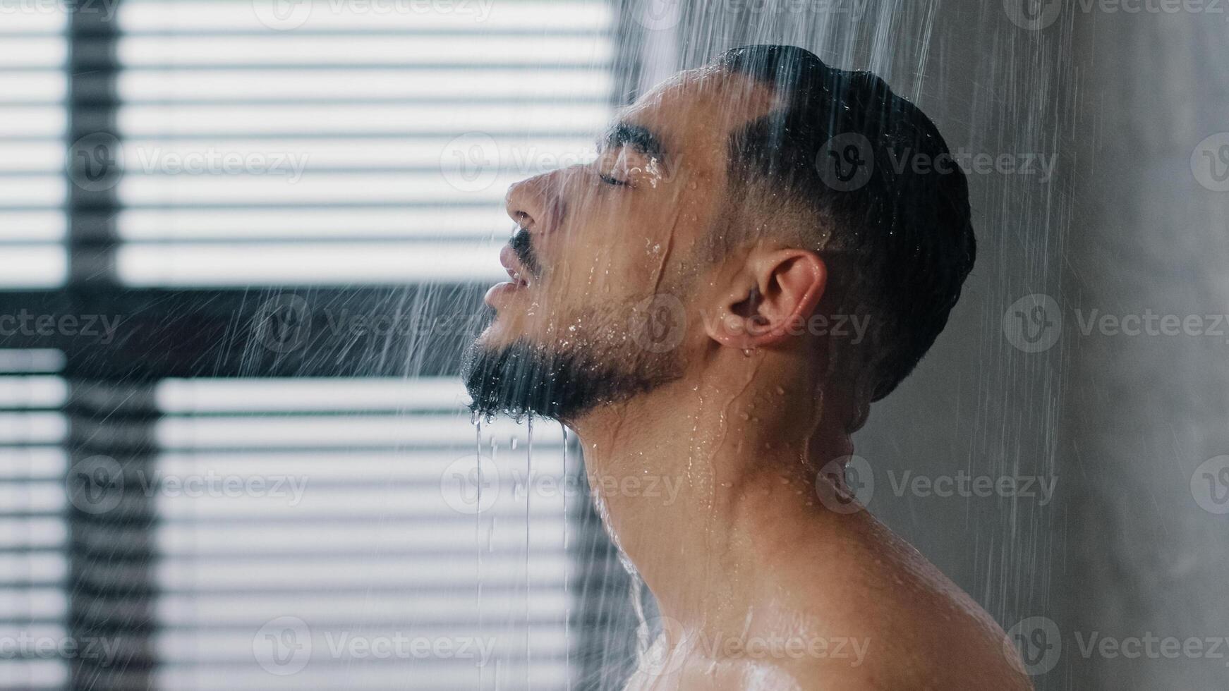 Side view closse up Arab relaxed man bearded guy naked male washes head hair face and beard in bathroom showering in at home in hotel after workout close up. Brunette rinsing shampoo and conditioner photo