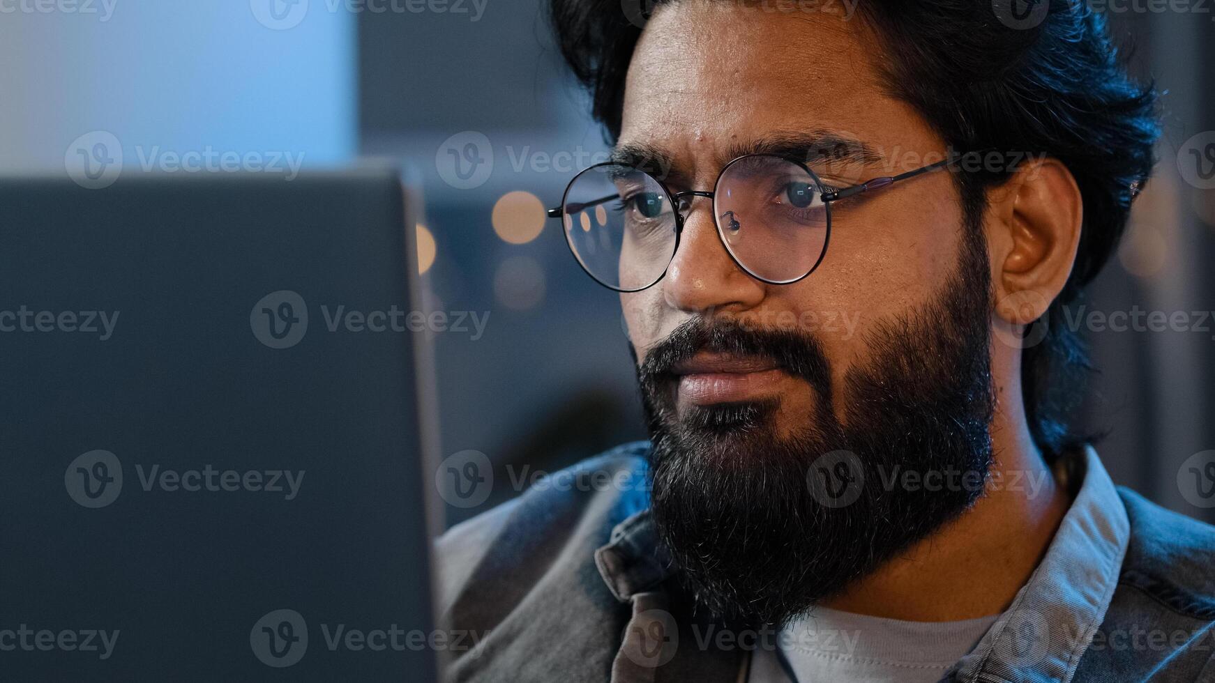 Close-up bearded male face reflection light from laptop monitor in glasses. Busy serious focused Arab Indian man looking at computer working freelance in evening playing game at night surfing in net photo