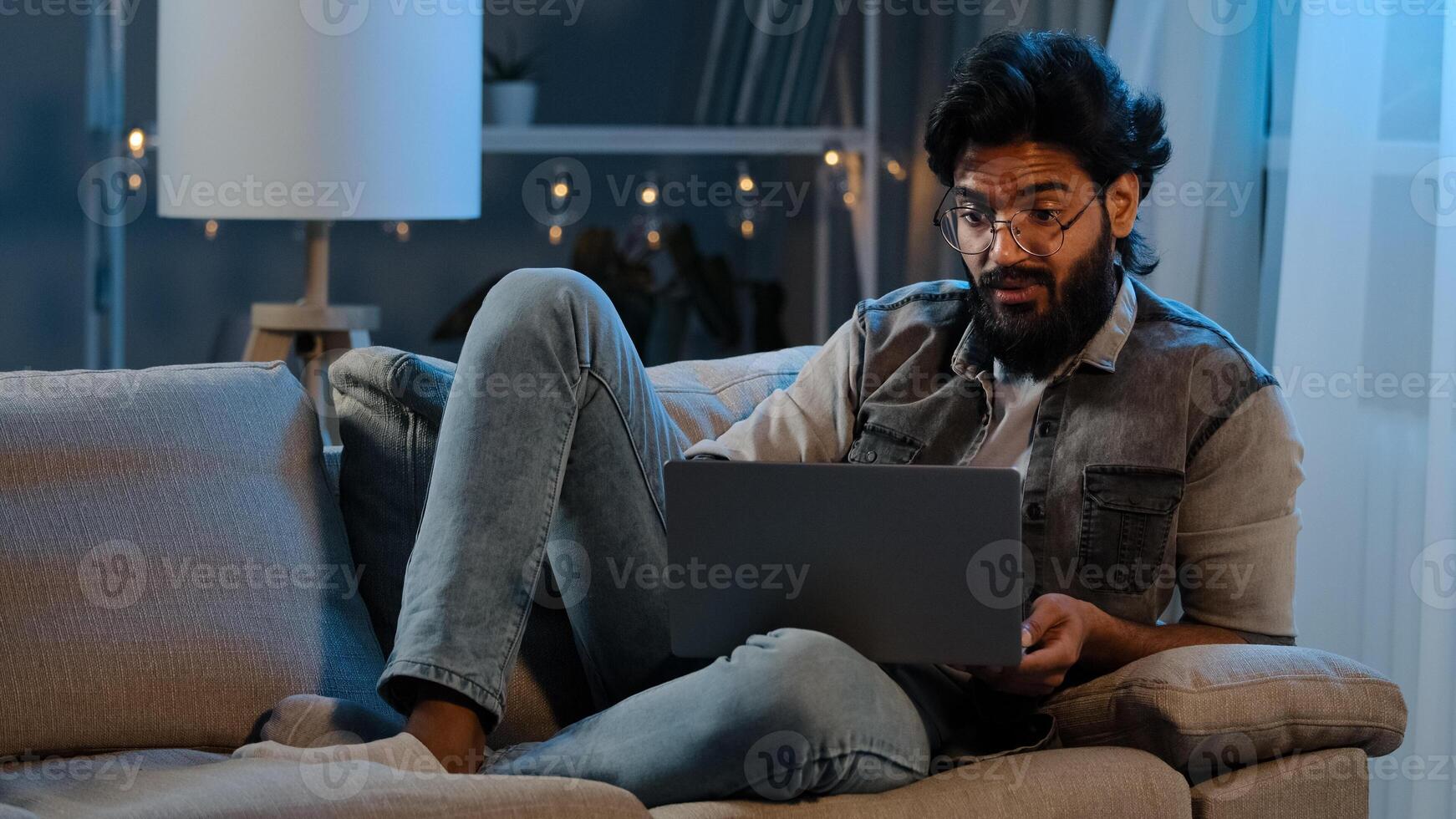 Businessman guy Arab Indian man bearded male user wearing glasses sitting on sofa at night late time with laptop reading bad news online trouble app job problems pondering solution looking at screen photo