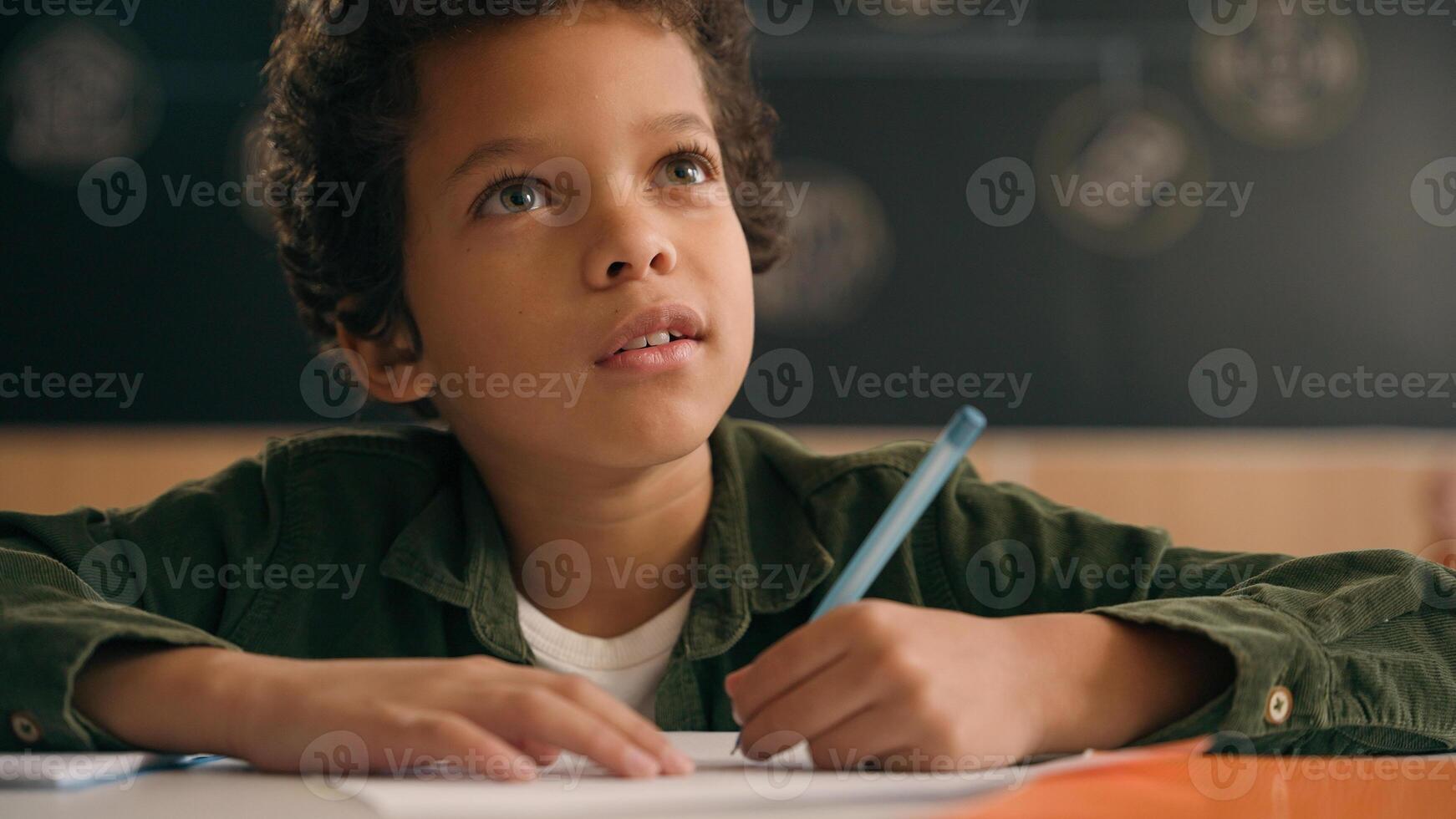 Little African American guy boy writing notes task class lesson primary school write in copybook kid schoolboy son child pupil student learning listen teacher studying education sitting at table desk photo