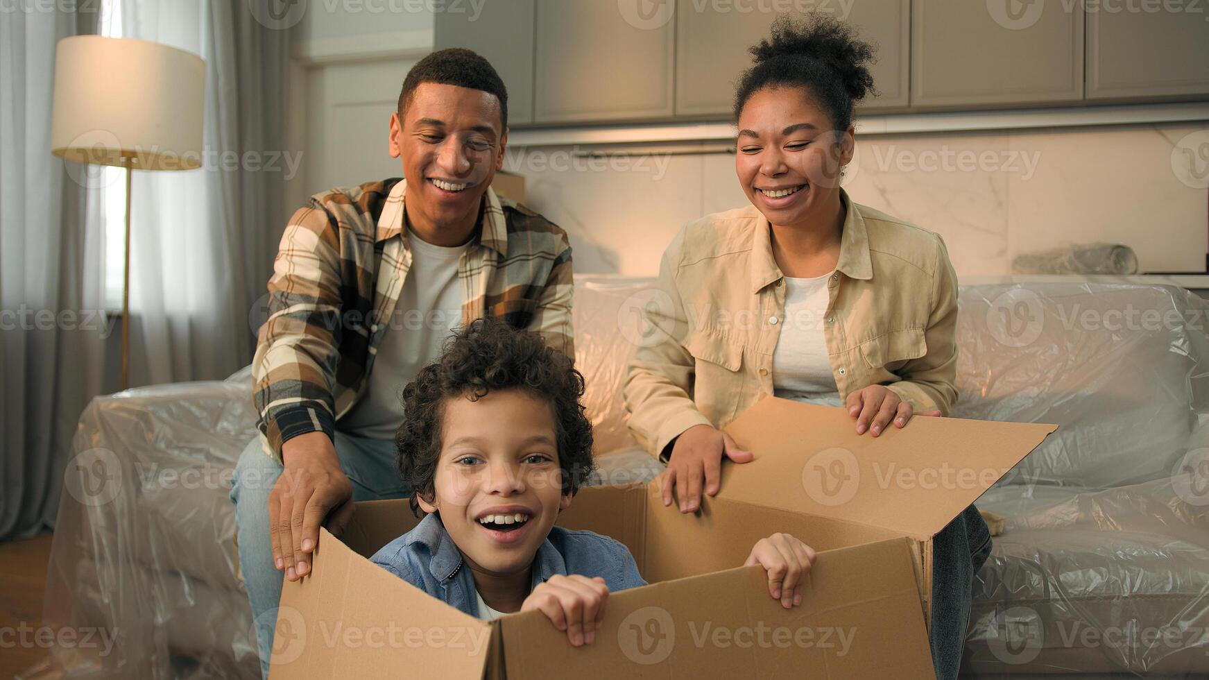 African American family moving in new home relocation happy parents unpacking parcel surprise funny joyful son little boy child jumping out of cardboard box playing game hide and seek kid having fun photo