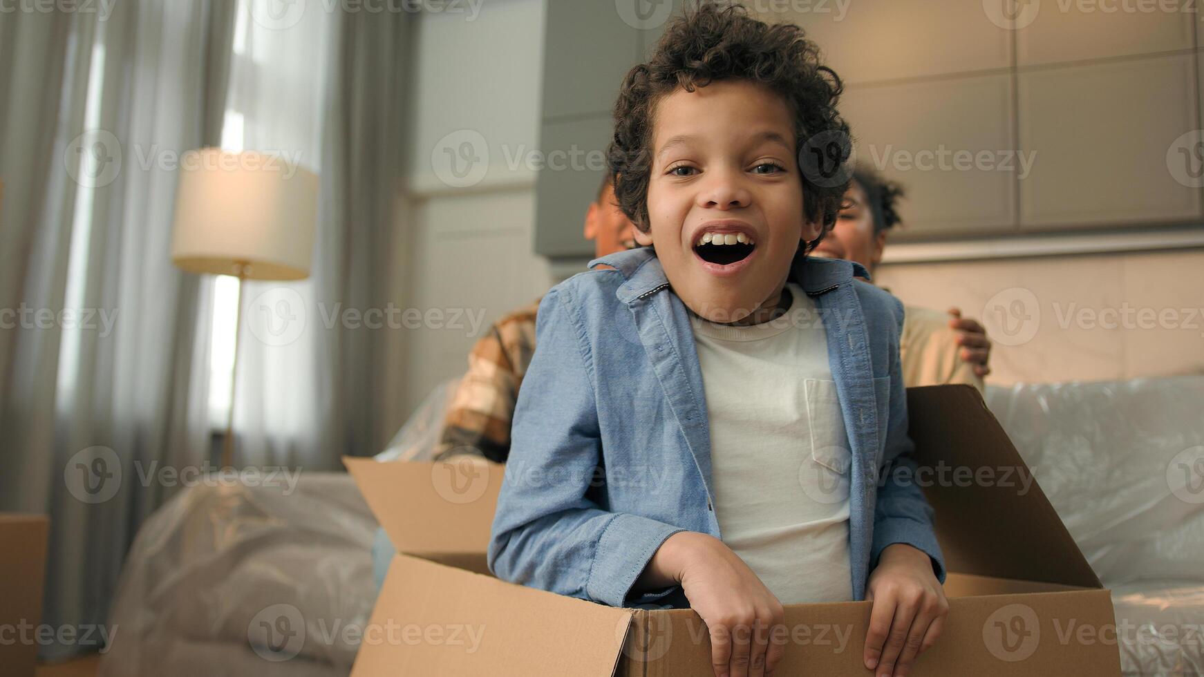 Happy family African American parents kid at home unpacking cardboard parcel new home relocation moving day surprise funny cheerful little boy child son jump out of box playing hide and seek game fun photo