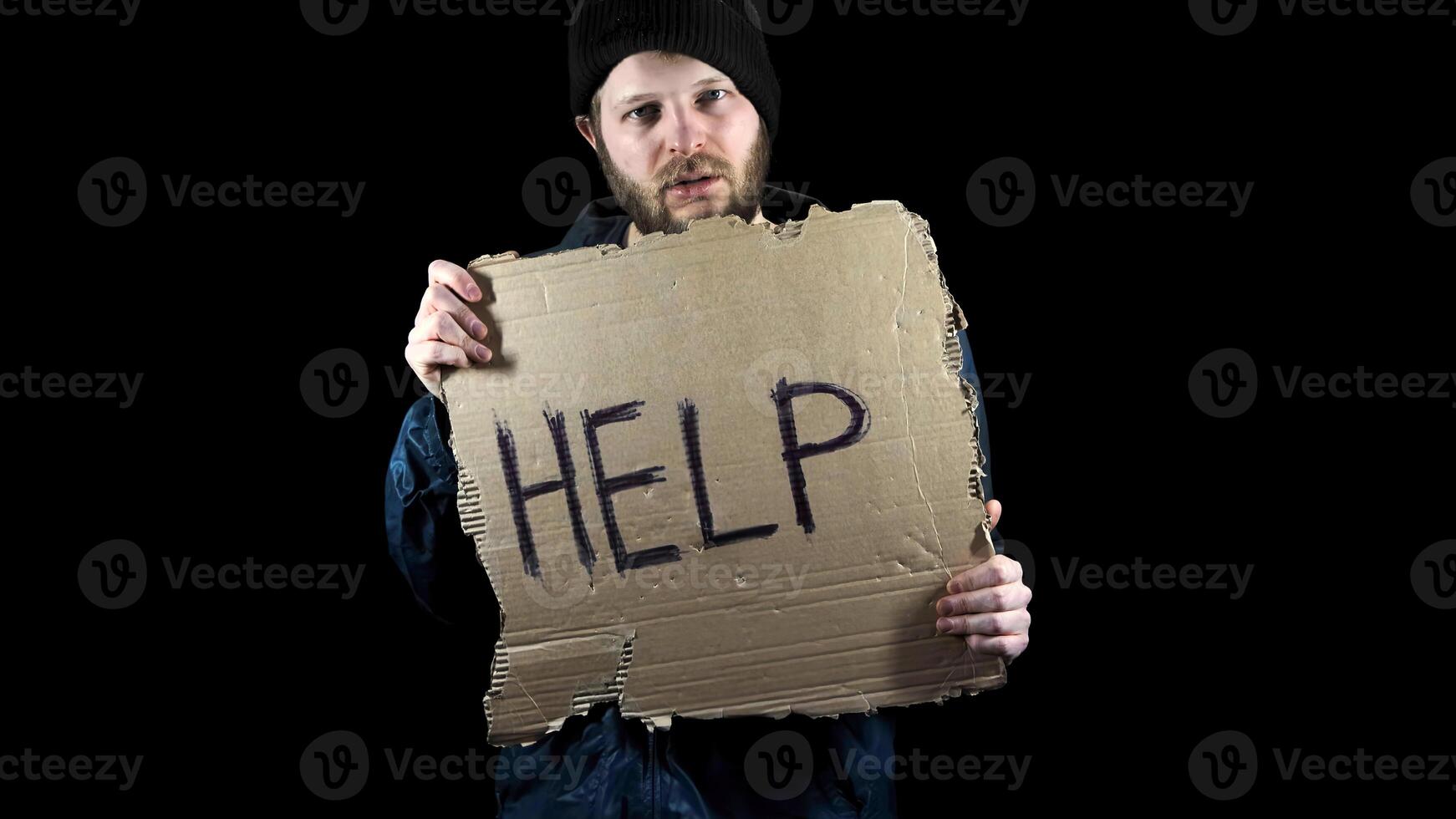 Homeless holds piece of cardboard with inscription Help. Refugee is looking for job. Male tramp in dirty clothes, black background. Poverty. Bearded fugitive beggar. Immigrant. Drunkenness photo