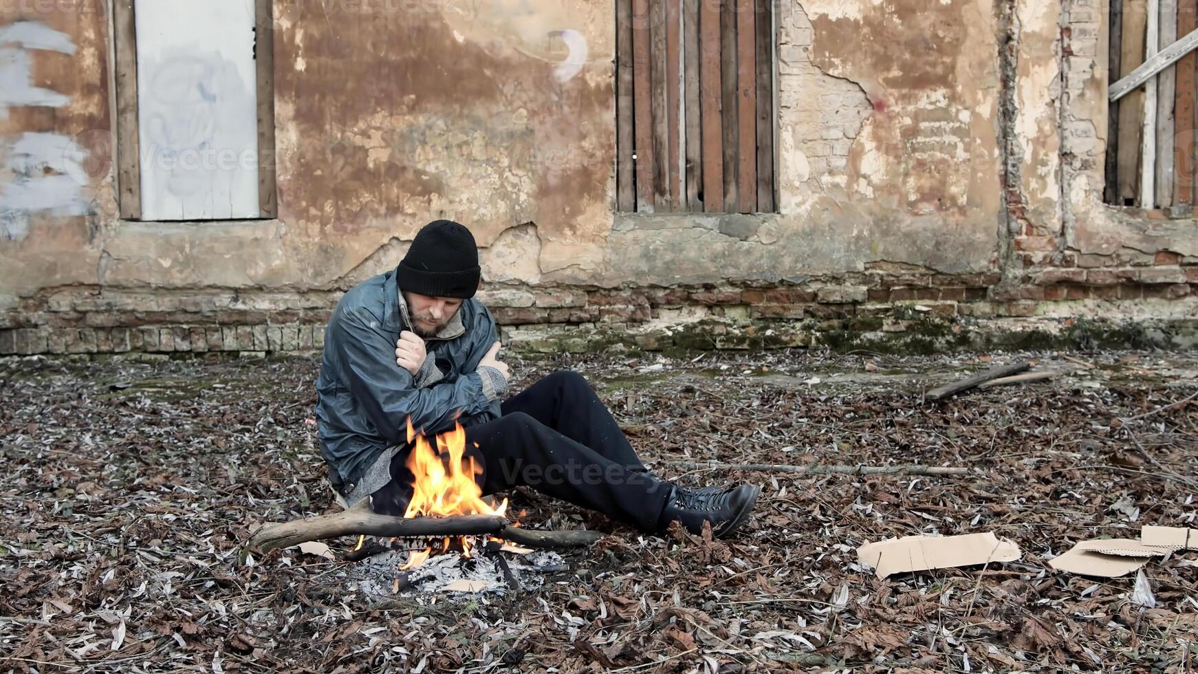 Homeless man sits on ground and pushes pieces of cardboard into fire to keep warm from cold. Tramp in dirty clothes and hat cap. Below poverty line. Illegal immigrant. Male refugee. Fugitive beggar photo