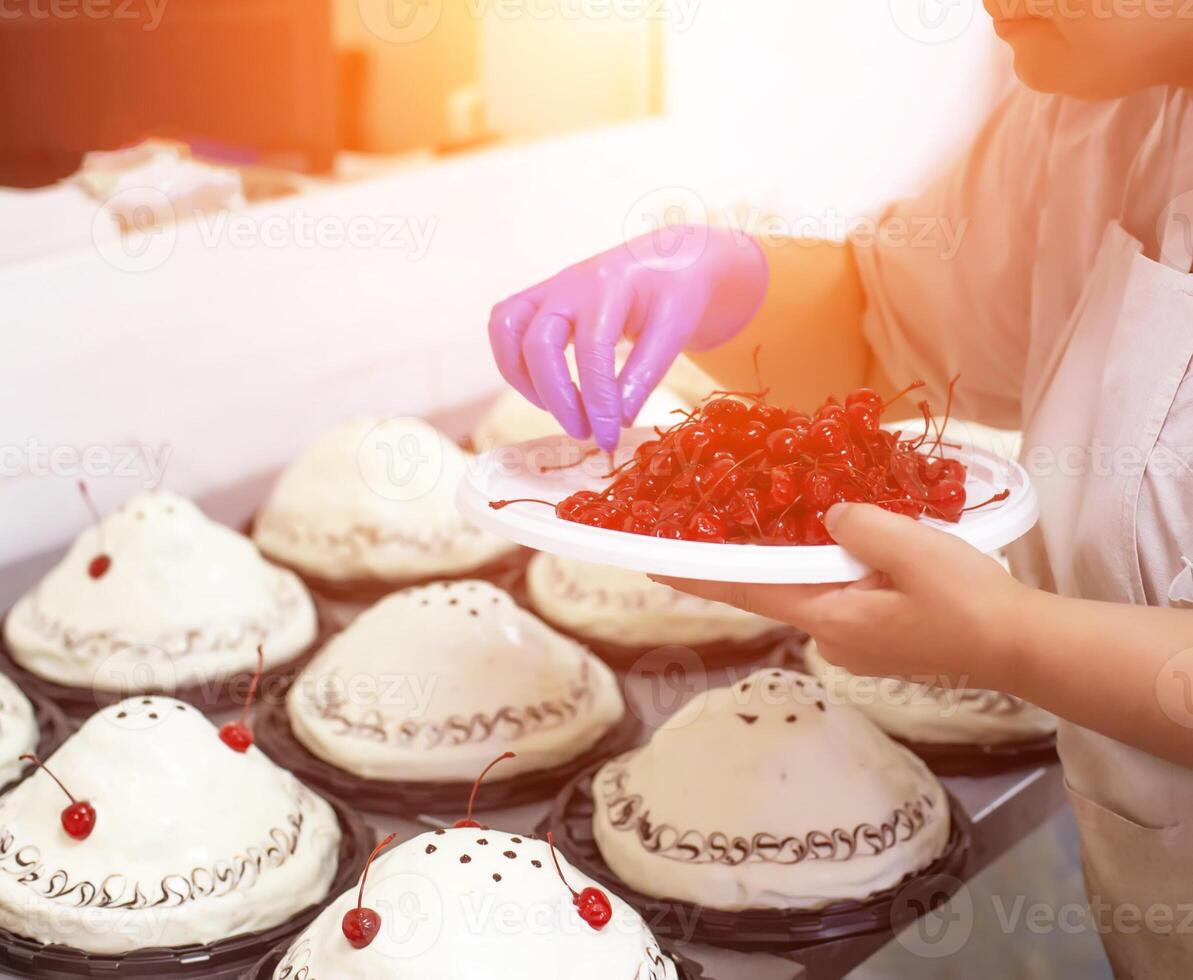 A chef decorates cakes in a confectionery factory with fresh cherry. Dessert mass production, background, industry, confectioner photo