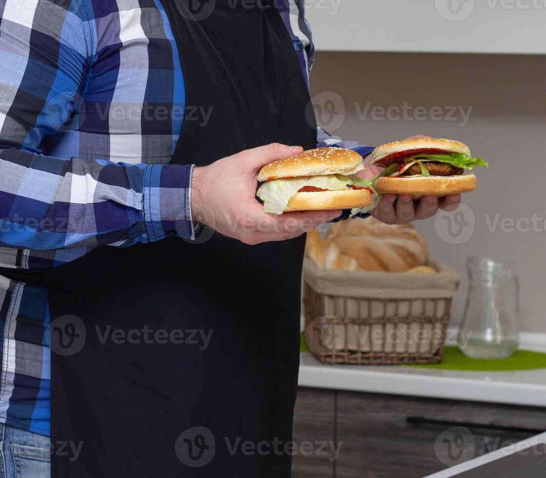 a man with a beard and in an apron holds burgers in his hands in the kitchen, background photo
