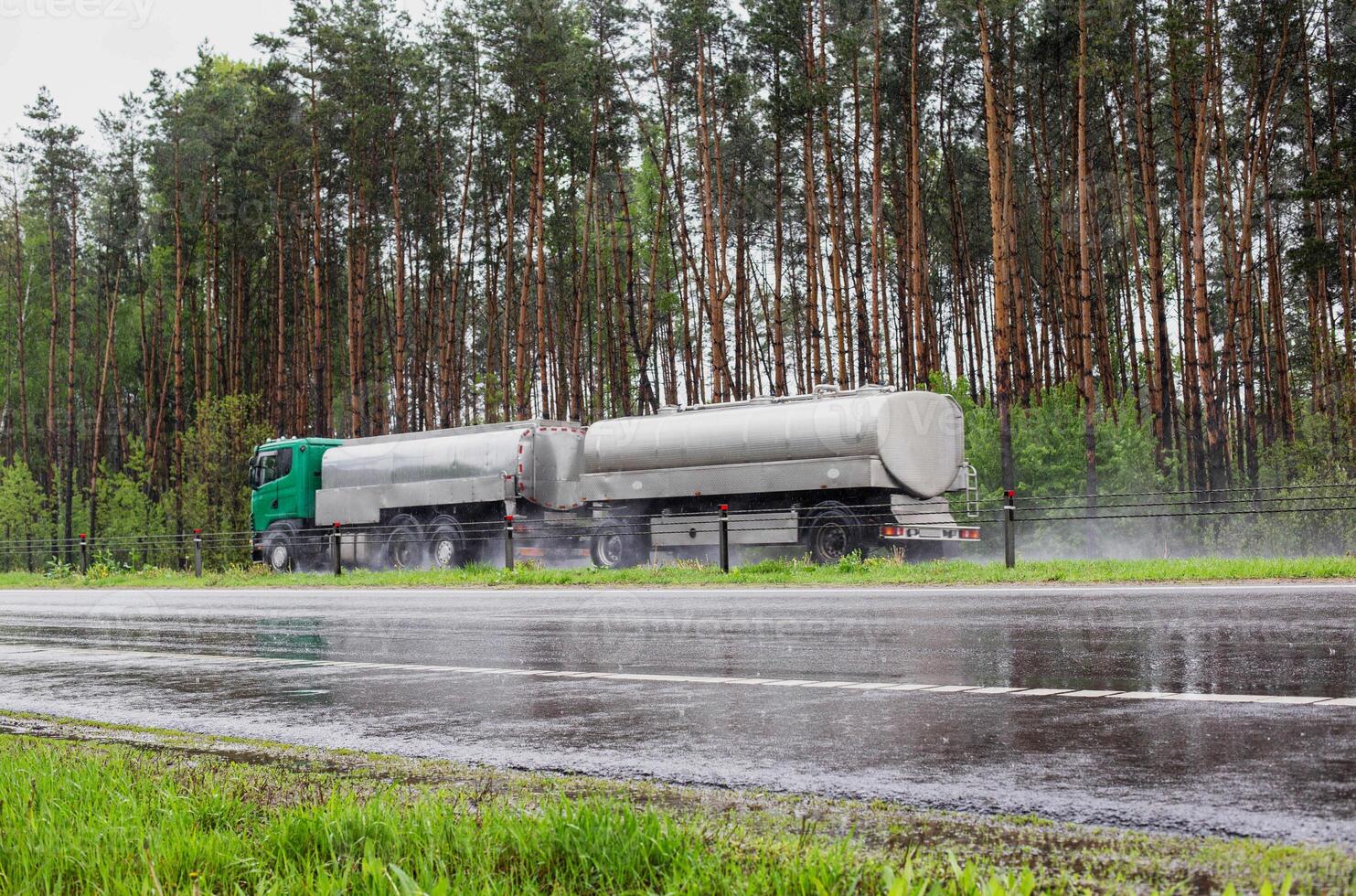 A truck with a tank semi-trailer transports liquid cargo on a slippery wet road in summer against the backdrop of a forest. Transportation of goods in bad weather. Sanctions photo