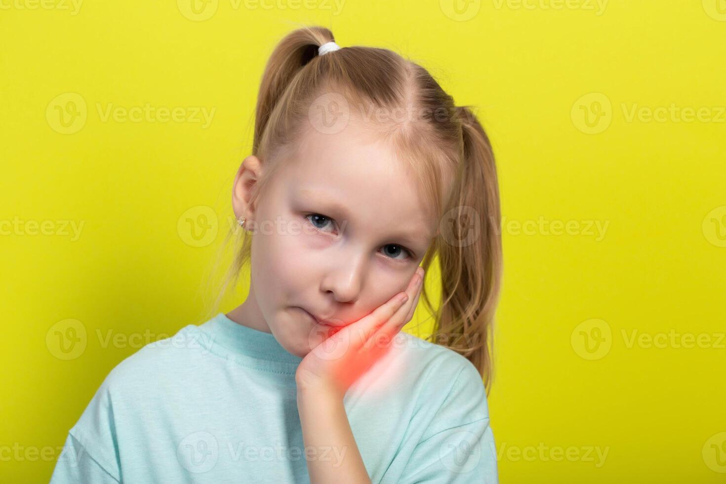 A beautiful little girl of seven years old holds her red, inflamed cheek with her hand. Concept of tooth pain in children, caries, gum inflammation and gingivitis. Yellow background, close-up. photo