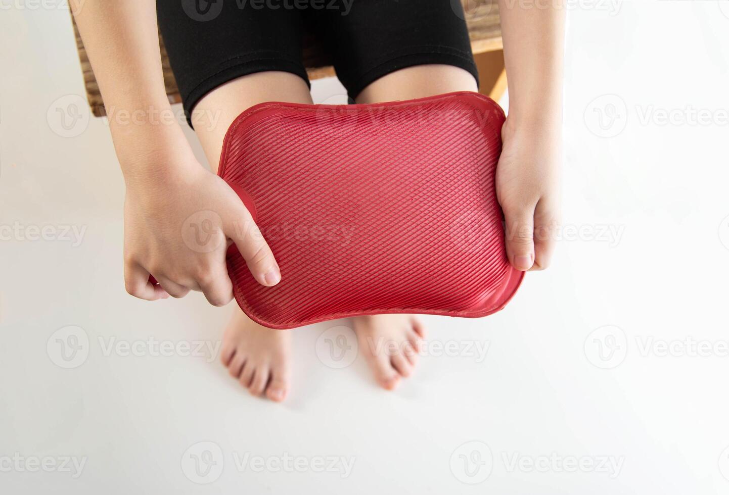 A little girl applies a red heating pad with hot water to her knee joint. Relieving pain with heat. Sprains in children, close-up photo