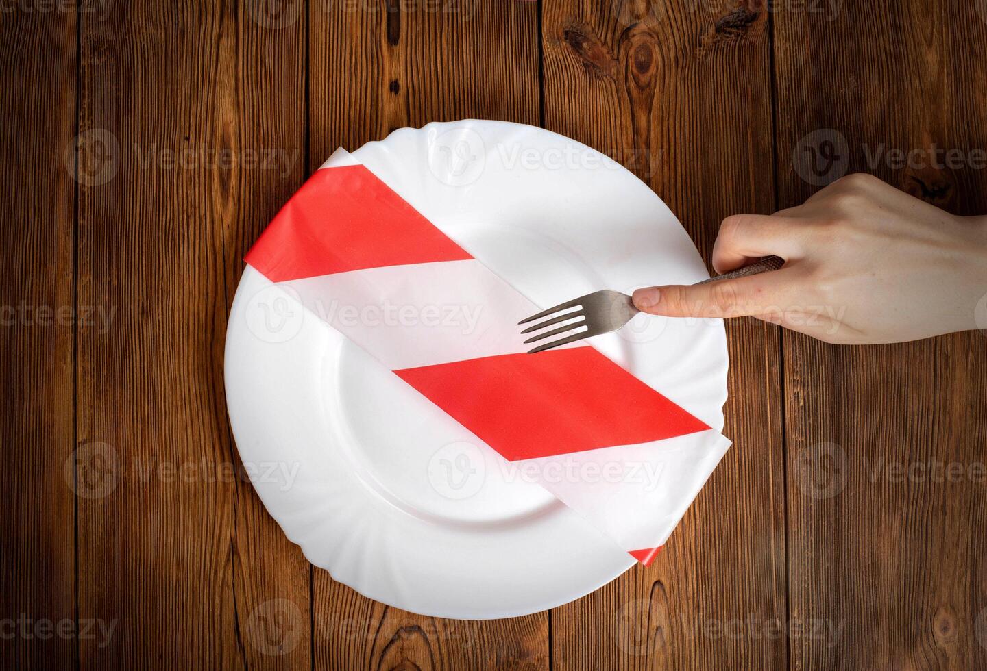 An empty plate with a forbidden white-red ribbon on a wooden background and a man's hand with a fork. The concept of therapeutic fasting and weight loss photo