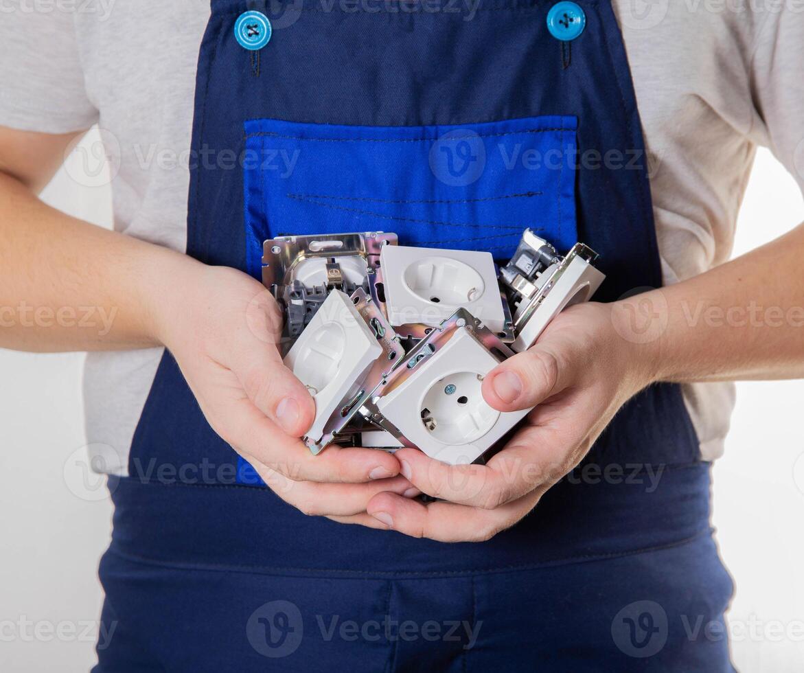 A man in blue overalls holds a lot of electrical sockets in his palms. The concept of electrician services, installation and connection of sockets, close-up photo