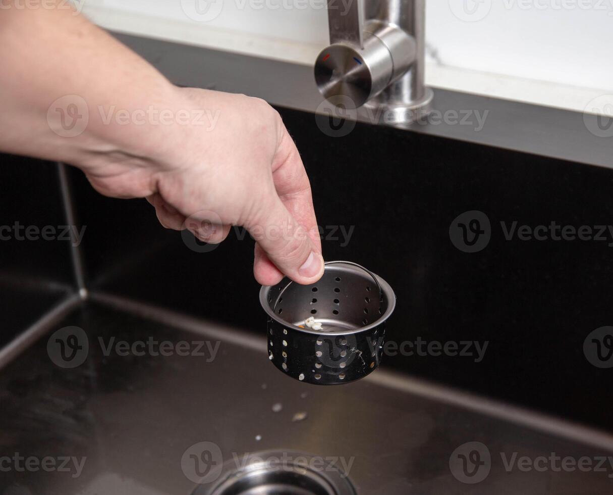 A man's hand removes a metal strainer from a kitchen sink drain. Cleaning the drain and pipes from clogging with food particles after washing dishes, close-up photo