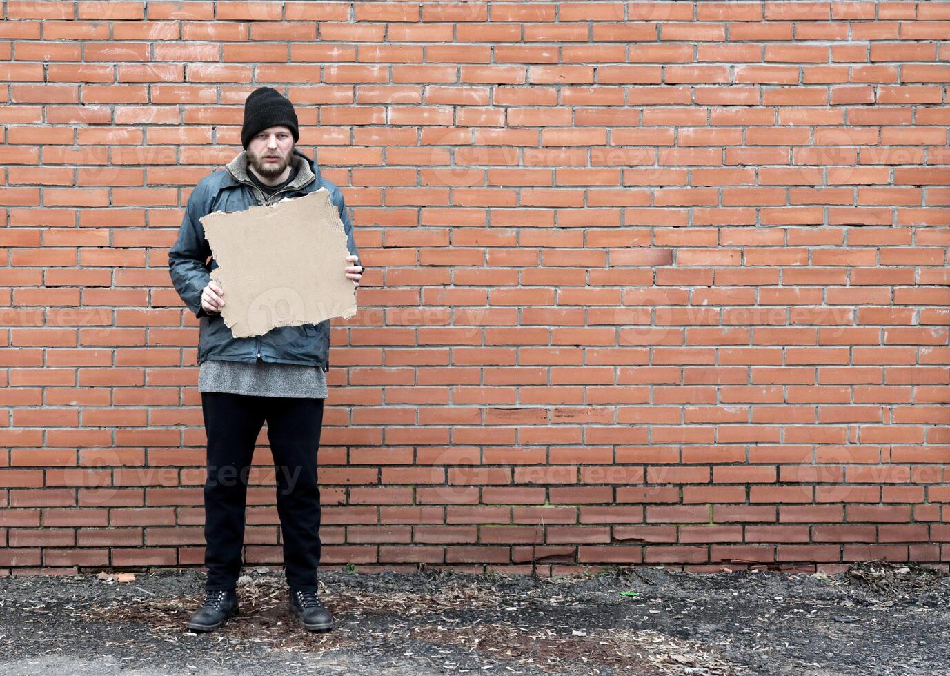 Homeless holds piece of cardboard. Refugee is at brick wall looking for job. Male tramp in dirty clothes. Below poverty line. Bearded fugitive beggar. Drunk immigrant. Copy space for text photo
