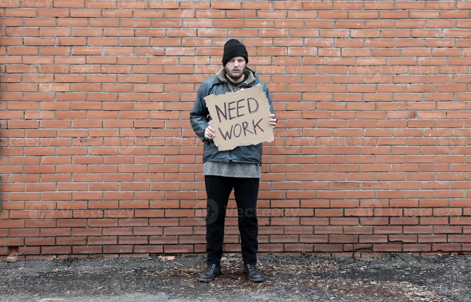 Homeless holds piece of cardboard with inscription Need Work. Refugee is at brick wall looking for job. Male tramp in dirty clothes. Below poverty line. Bearded fugitive beggar. Immigrant. Drunkenness photo