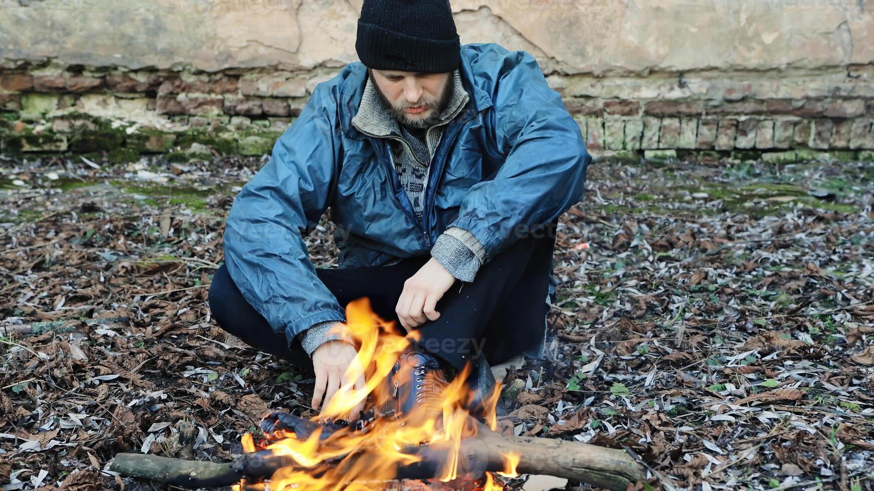 Homeless man sitting and warming himself by the fire from cold. Tramp in dirty clothes and hat cap. Below poverty line. Illegal immigrant. Male refugee. Fugitive beggar photo