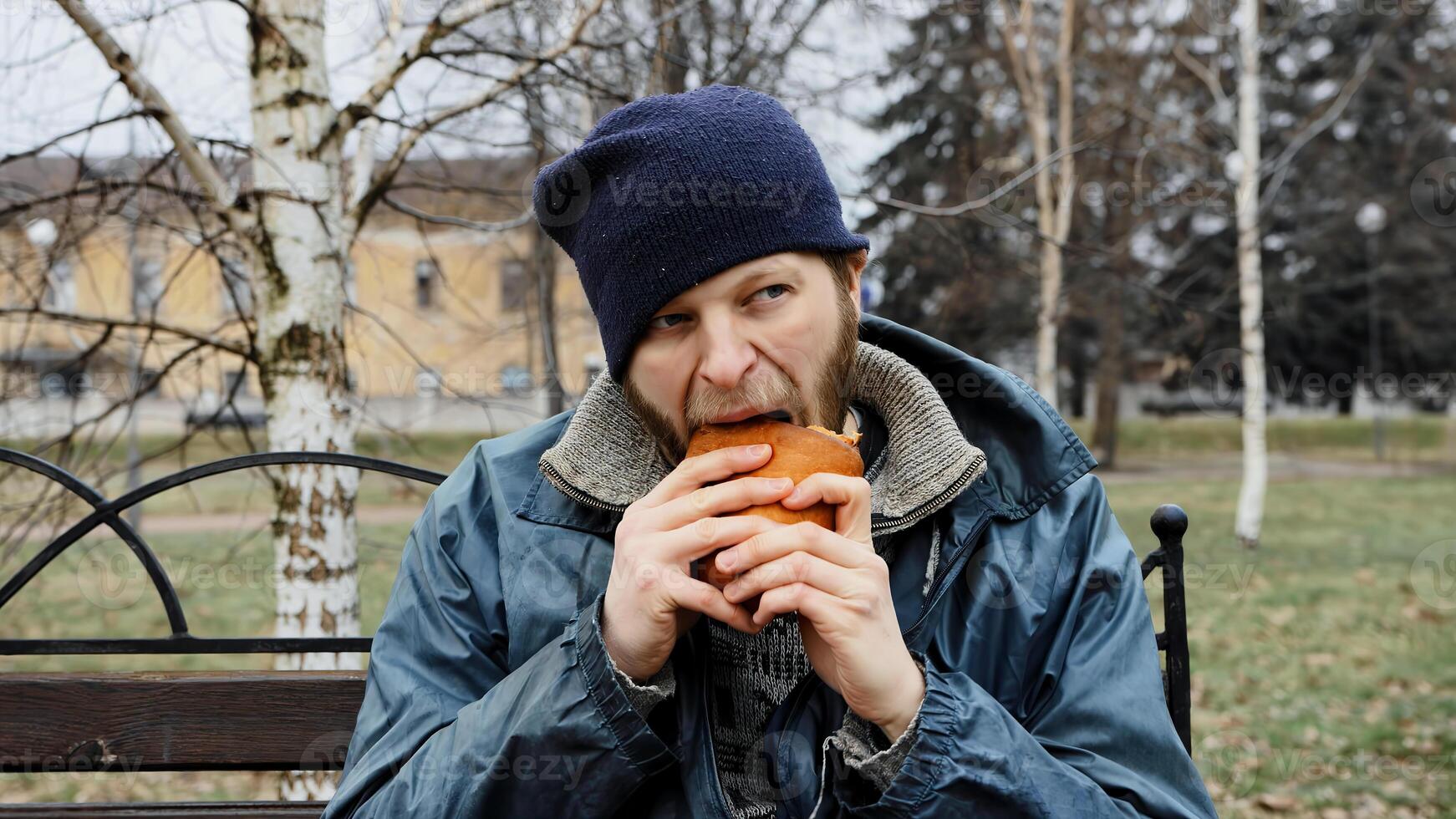 Hungry bearded homeless man sits on a bench and eats a bun in a city park. Below poverty line. Male tramp in dirty clothes with cap and hat. Beggar. Illegal immigrant. Drunkenness, alcoholism photo