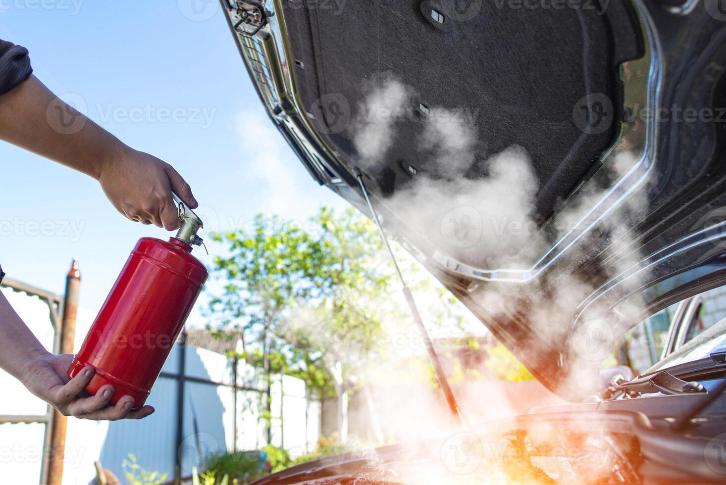 The driver of the car directs a powder fire extinguisher at the engine compartment of the car in which there is fire and smoke. Car fire, background. Electrical short circuit photo