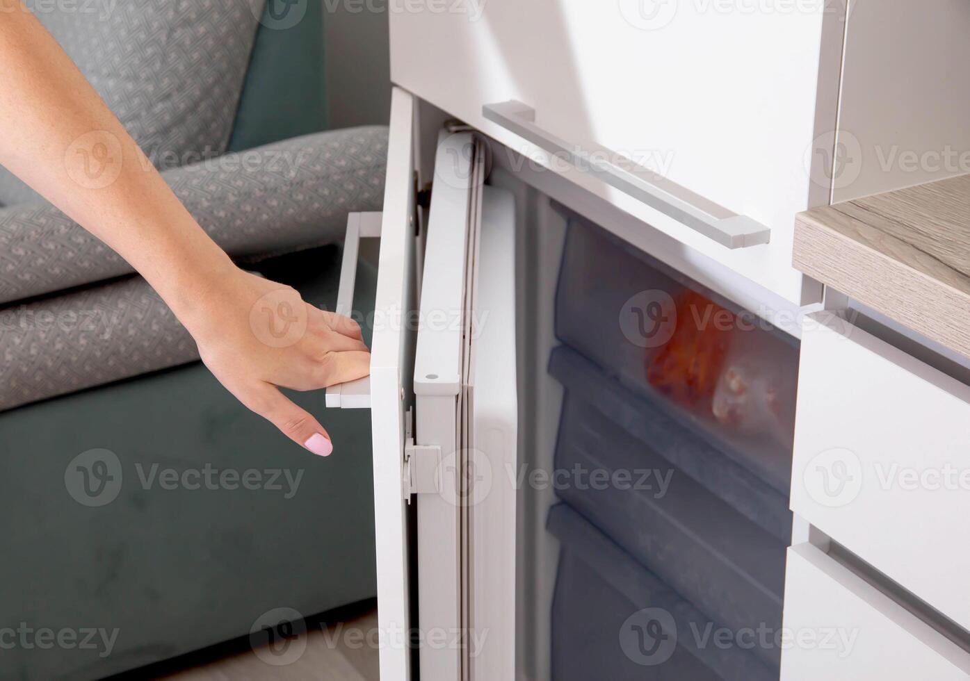 A woman's hand opens the door of the built-in freezer in a modern white kitchen, close-up photo