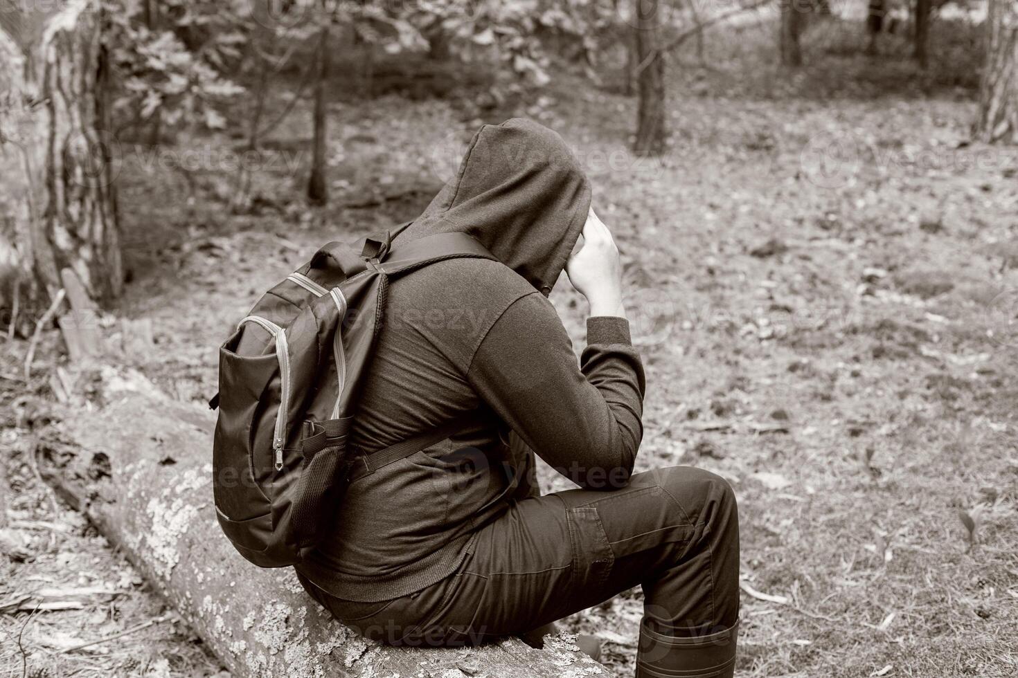 A man with a backpack in a hood sits on a fallen tree in the middle of a clearing in the forest. Survival in the wild. Dehydration of the body. Mushroom picker lost in the forest. Black and white photo