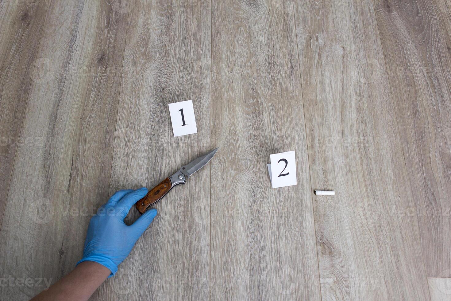 Criminalist in blue glove raises a collapsible knife from the floor, a cigarette butt on a wooden background, a close-up, an investigation photo