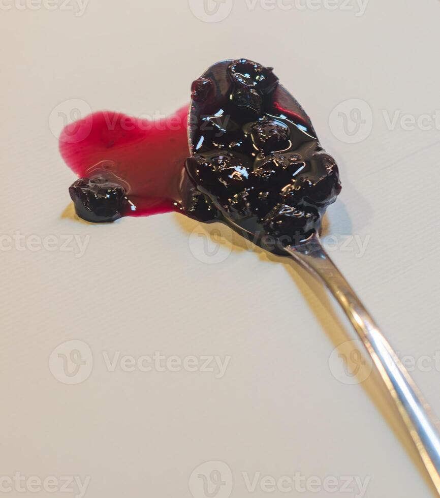 Spoon with a heart-shaped spill of dark red jam, with berries on a light surface. High quality photo