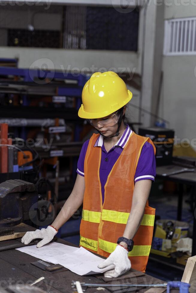 Asian woman technician Engineer in vest and helmet look and checking paper work at industrial workshop. photo