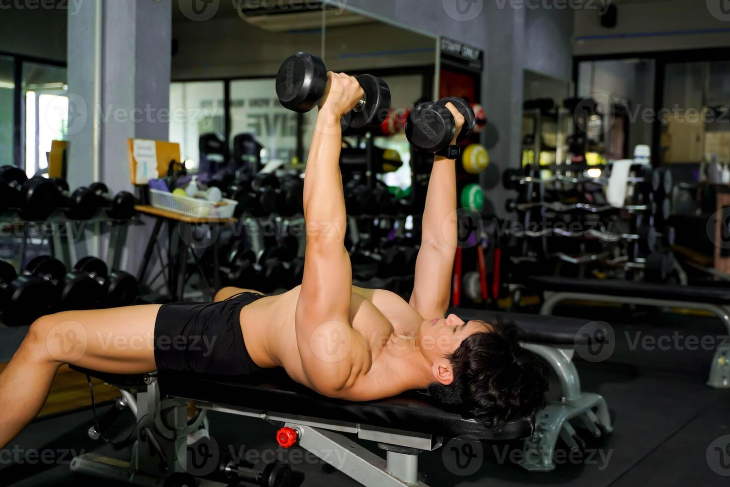 Closeup Asian handsome man with perfect body playing dumbbells' weight training in the lying position to build shoulder and chest muscles at fitness center. photo