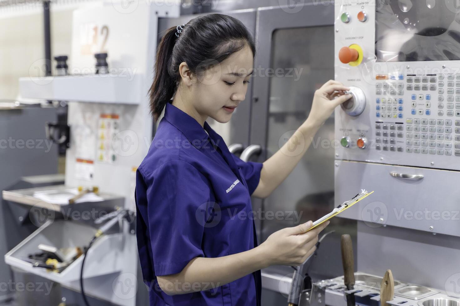 Asian woman industrial engineer wearing uniform looking at a clipboard and checking automated machinery in industrial workshop. photo
