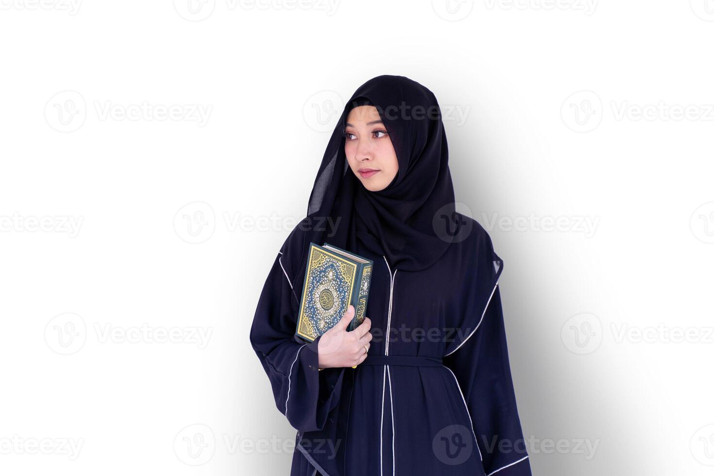 A beautiful Muslim student wearing abaya niqab traditional clothes standing and hold Quran on white wall background. Arabic texts is meaning Glorious Quran in English. photo