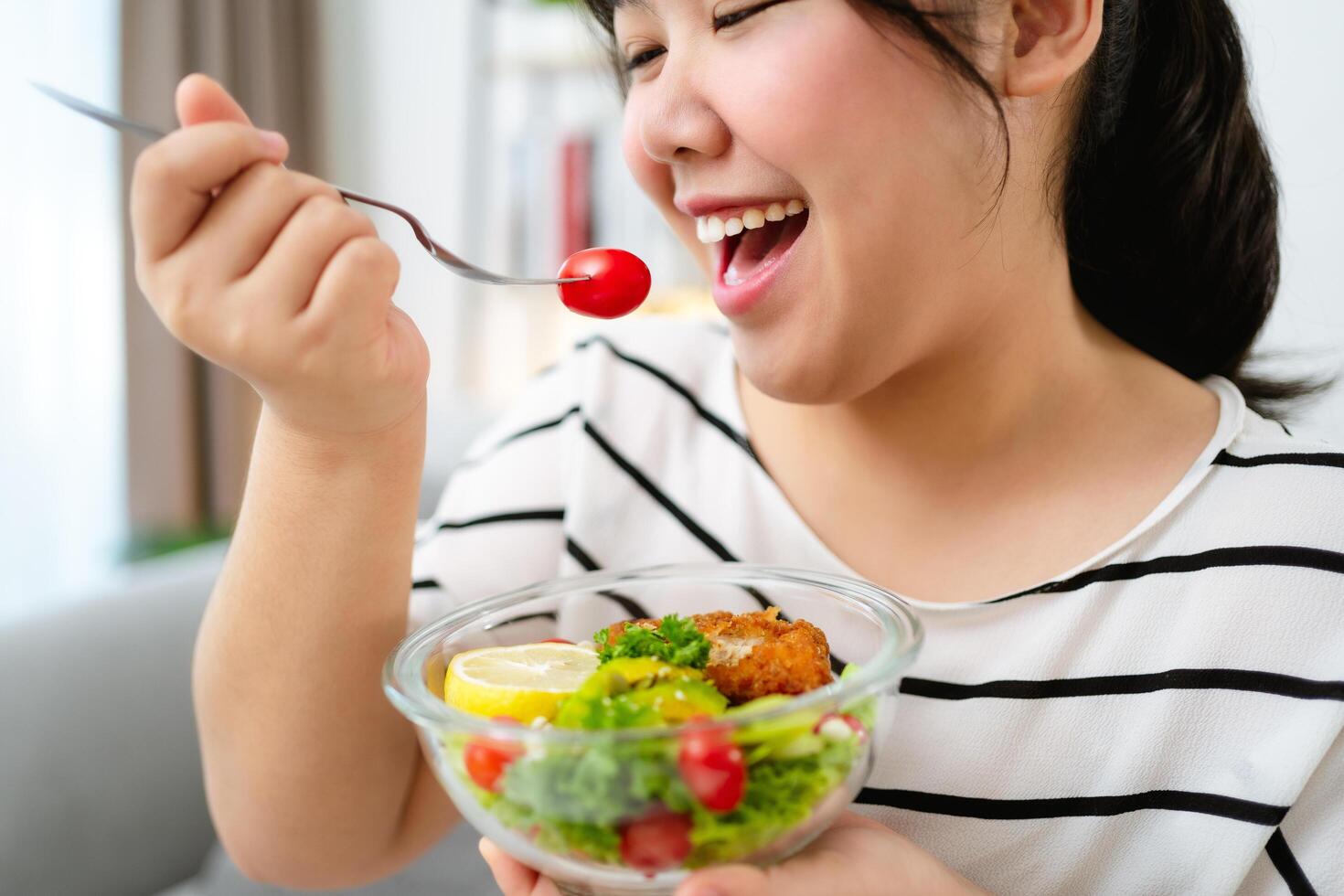 Beautiful Asian woman eating salad, holding a glass bowl and using a fork to pick up a tomato to eat photo
