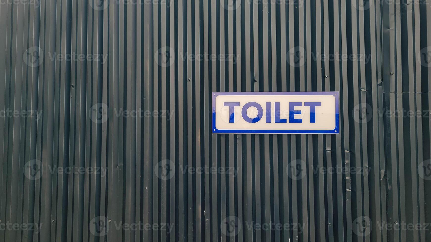 Photo of a toilet sign against a dark corrugated wall background. The sign, with bold and clear lettering, indicates the location of the toilet or restroom.