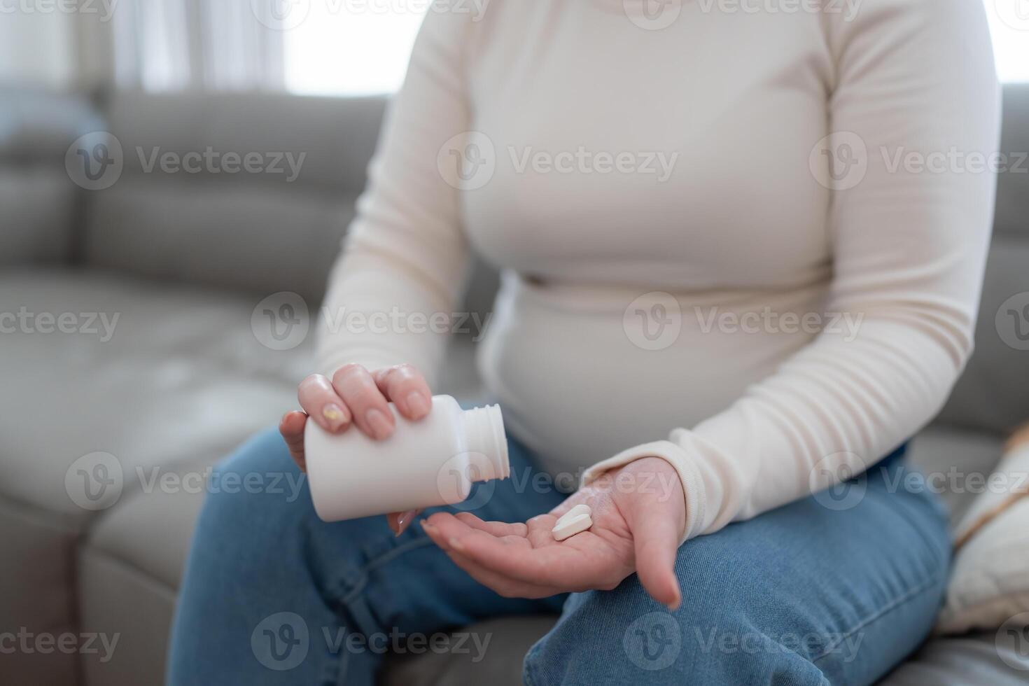 Unrecognizable pregnant woman holding jar with pills taking vitamins treating sickness during pregnancy sitting on sofa. Close up photo