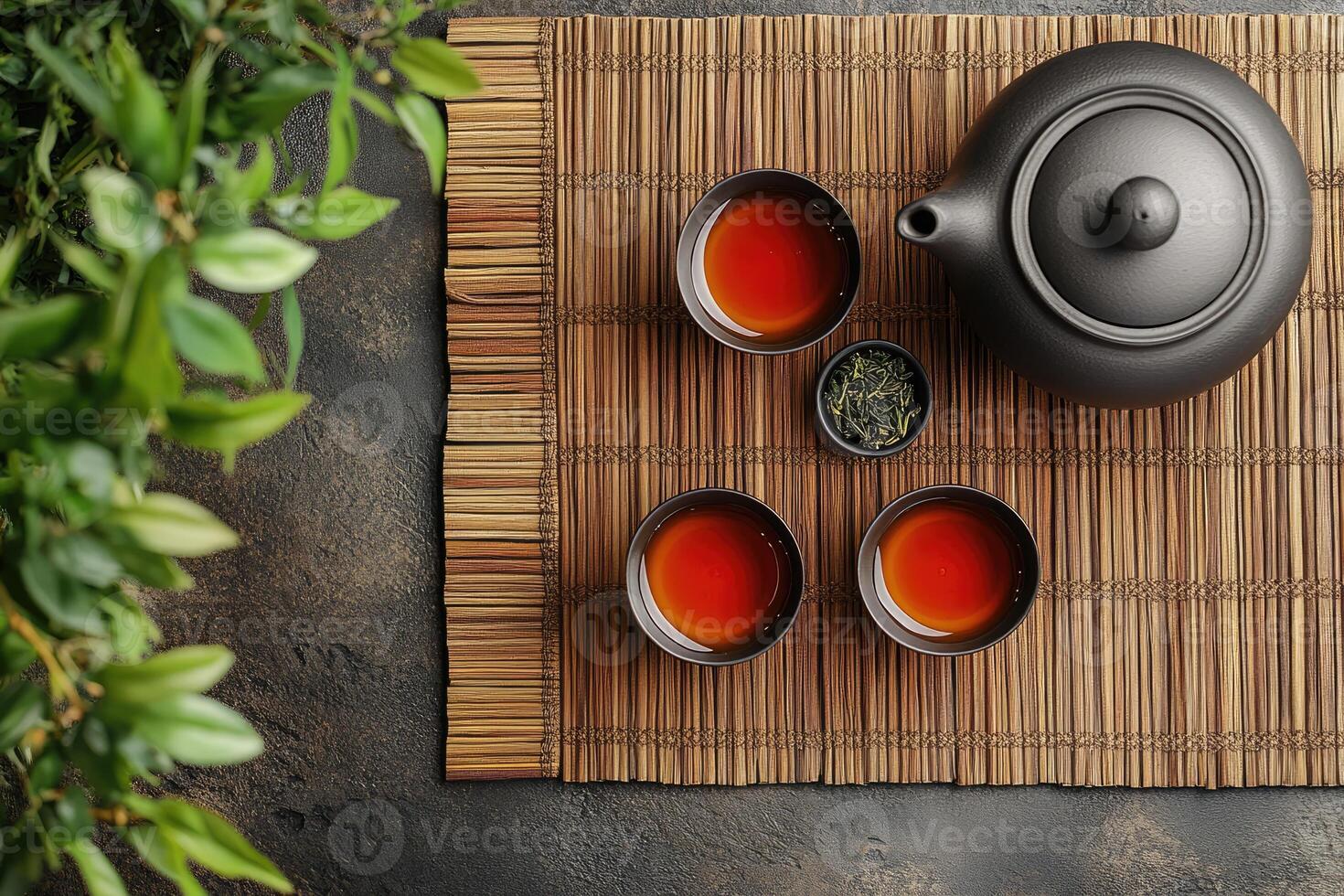 Black ceramic teapot and teacups standing on bamboo mat with green tea leaves near plant photo