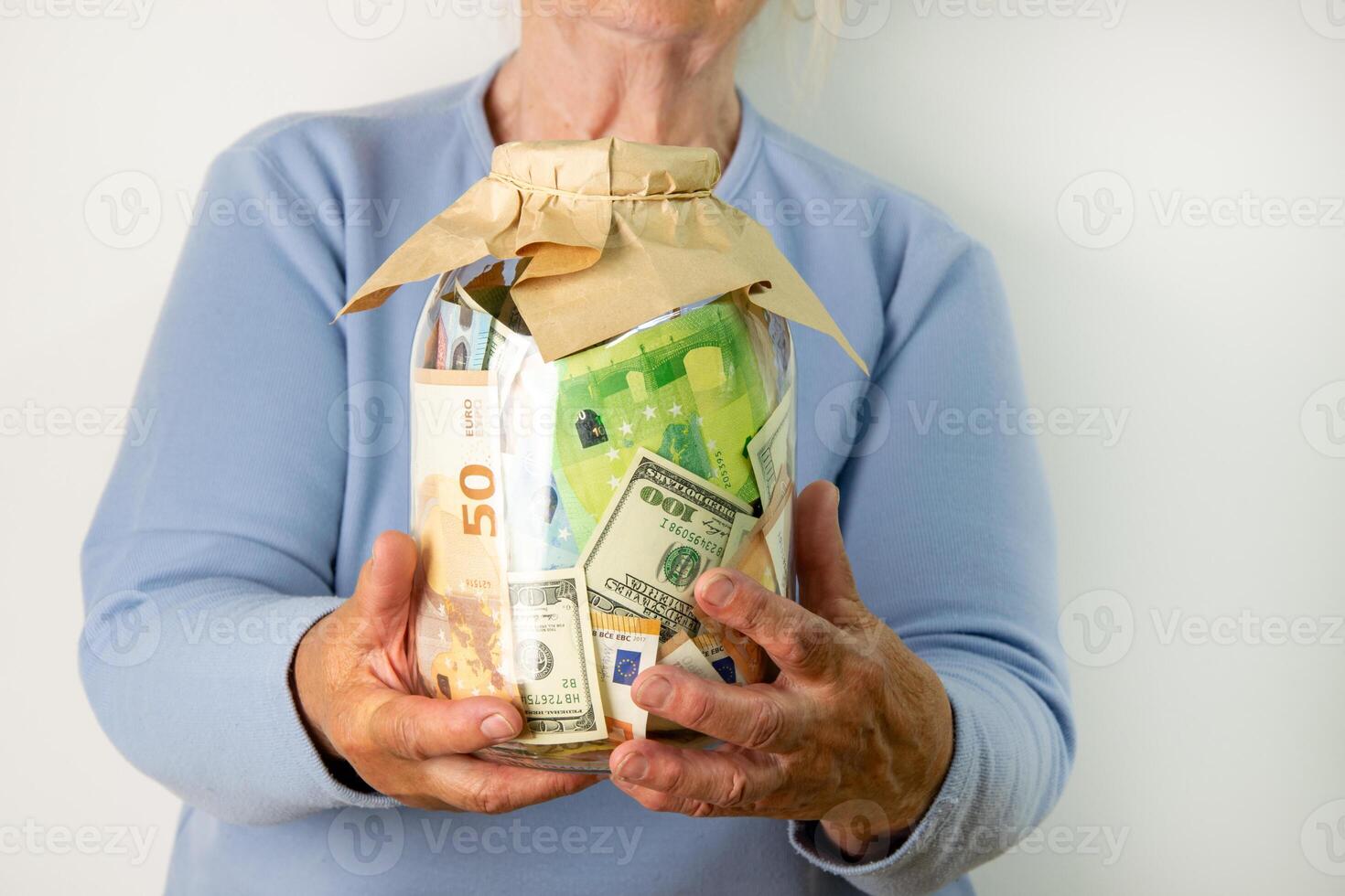 A senior woman in a blue sweater holds a glass jar filled with various US dollar and euro bills photo