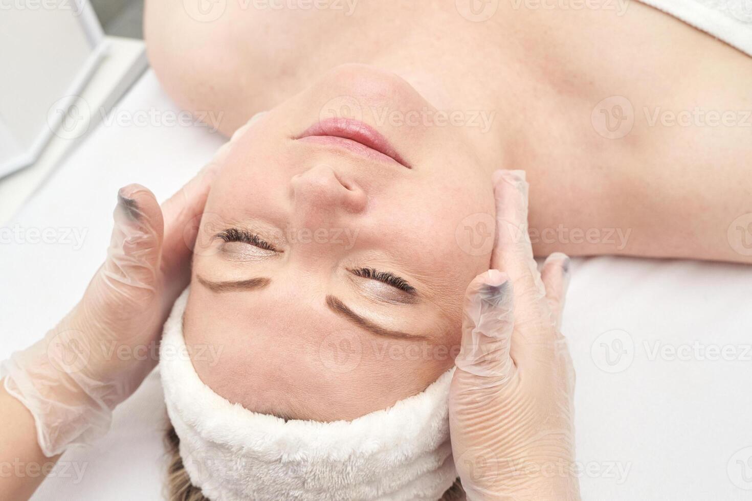 Young caucasian female receiving relaxing facial massage at spa with therapist in white gloves. photo