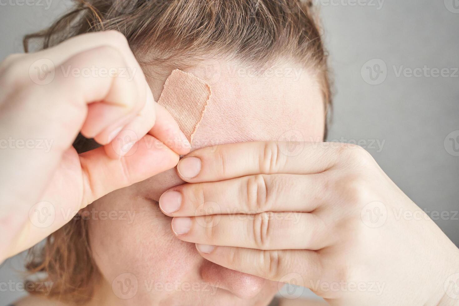 Adult caucasian female applying bandage to forehead with close up view of hands and hair in neutral setting. photo
