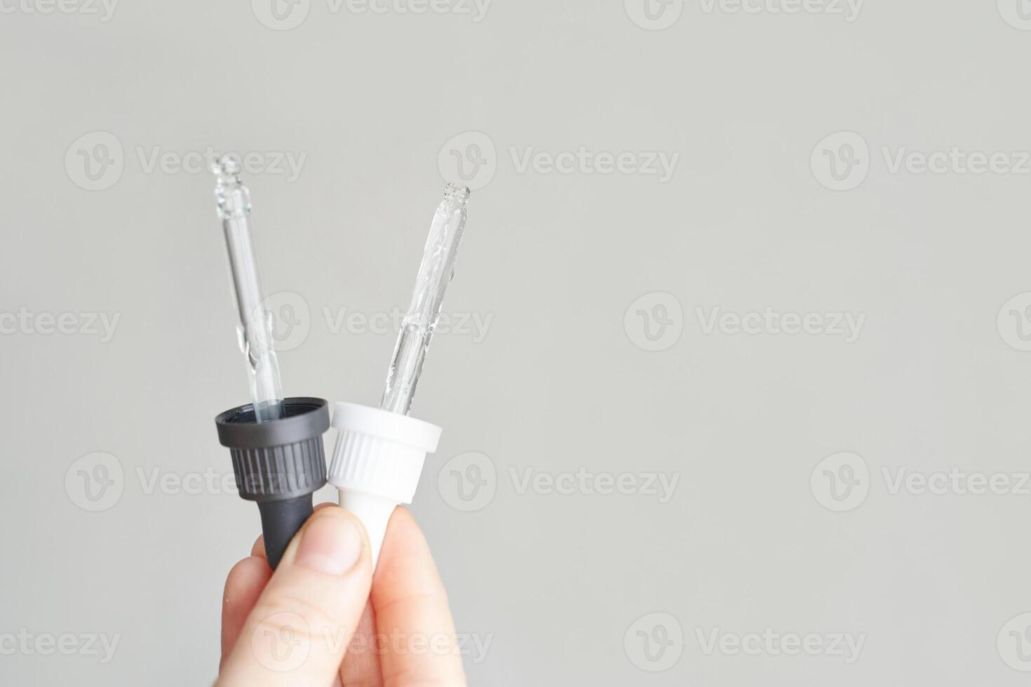 Close up of two glass droppers with white and black rubber tops held in hand against neutral background. photo