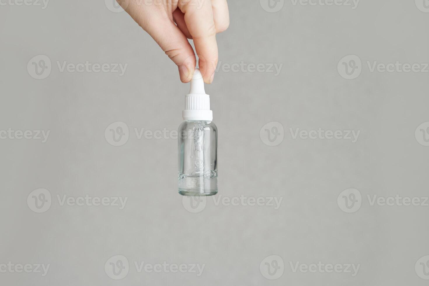 Hand holding clear dropper bottle on gray background for skincare or essential oils display. photo