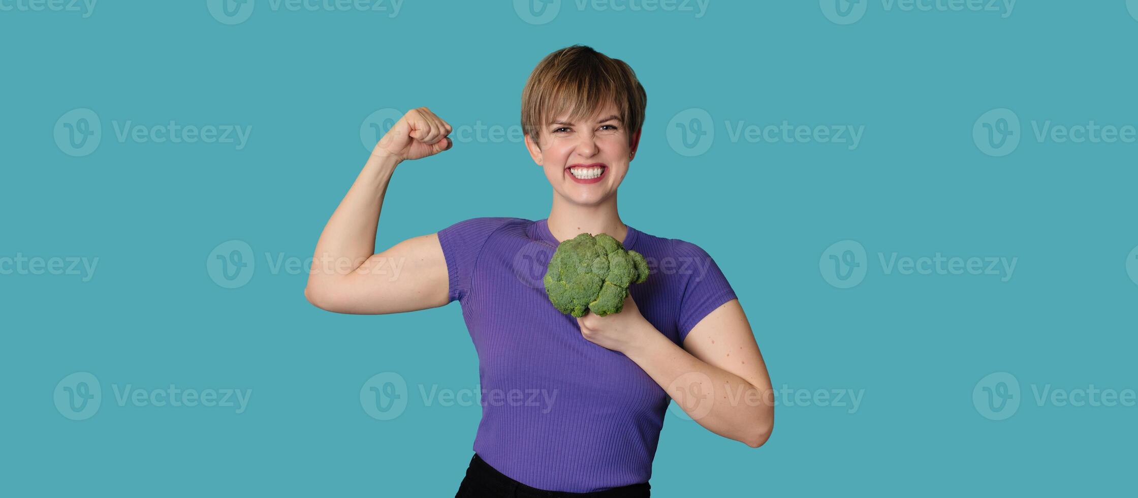 Smiling Caucasian girl with fresh broccoli in one hand and showing biceps with the other. Panoramic banner, free space. photo