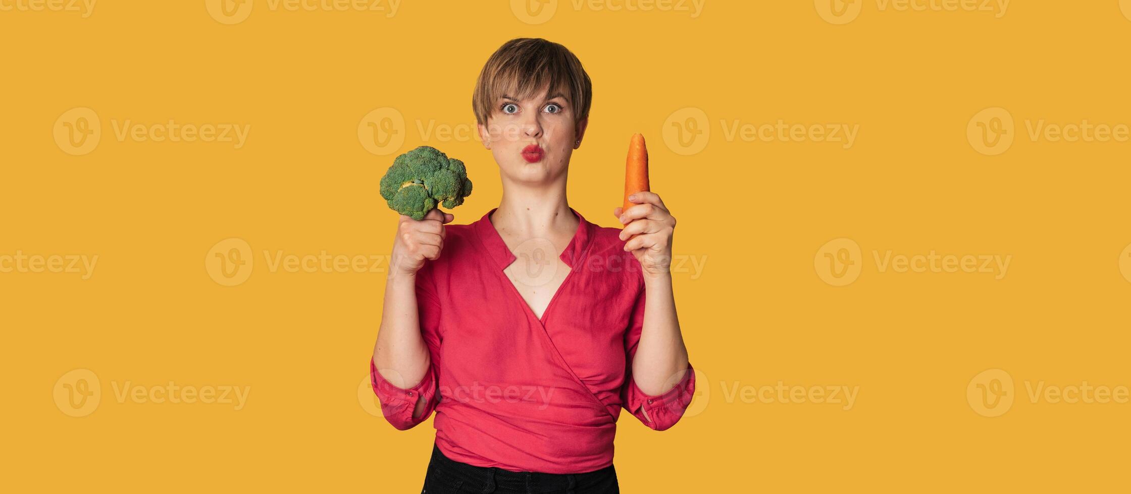 A studio shot of a smiling young woman with fresh broccoli and carrots in her hands on a yellow background. The concept of healthy eating. A place for your text. The panorama. photo