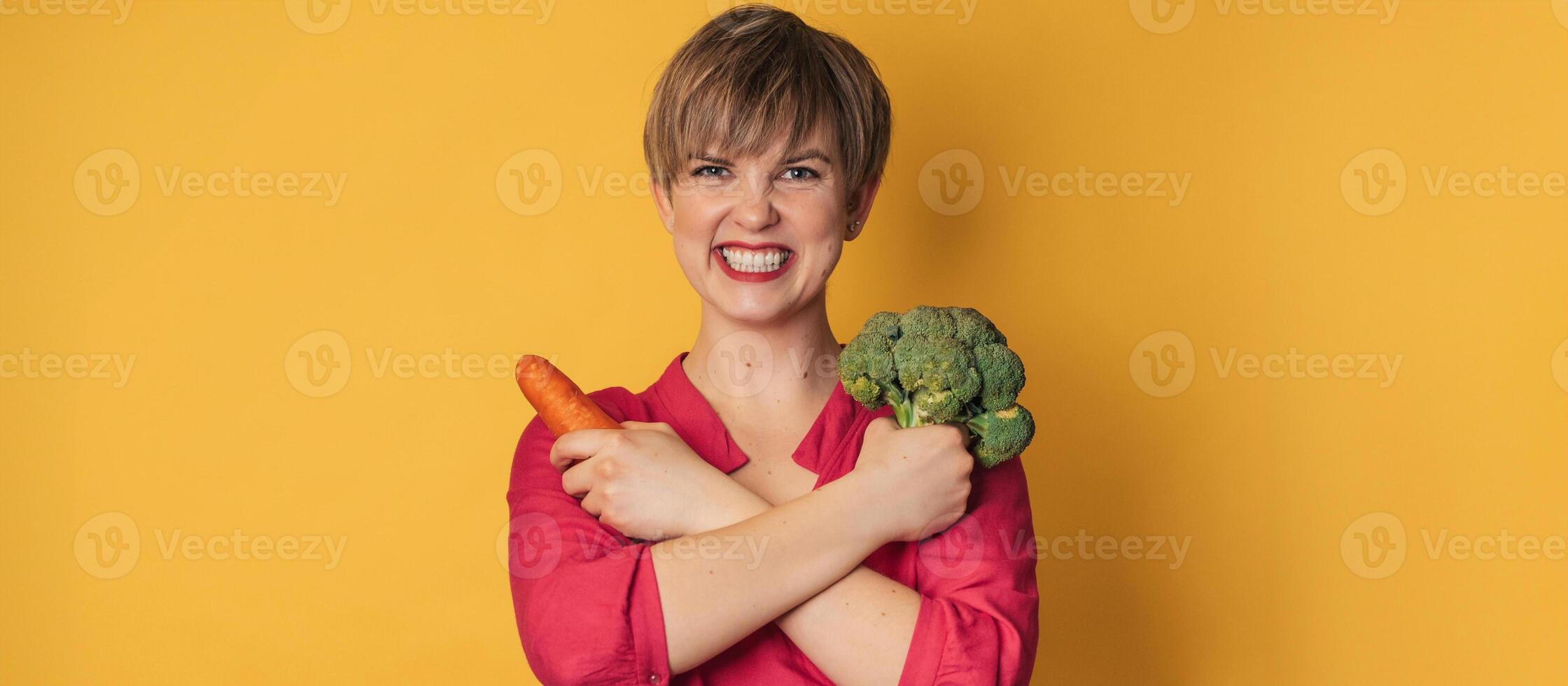 A studio shot of a smiling young woman with fresh broccoli and carrots in her hands on a yellow background. close-up.. The concept of healthy eating. A place for your text. The panorama. photo
