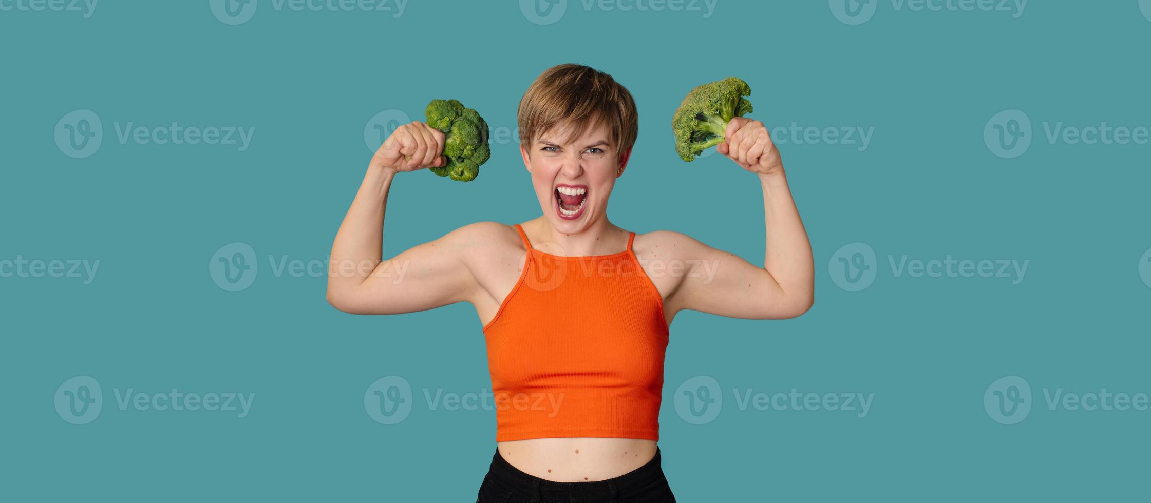 An expressive portrait of a young woman with broccoli as dumbbells on a blue background. Healthy diet, healthy lifestyle. A panoramic banner with a copy of the space. photo