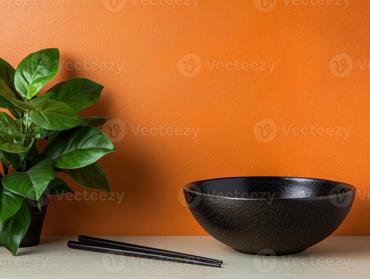 Black bowl and chopsticks on a table with a green plant against an orange wall photo