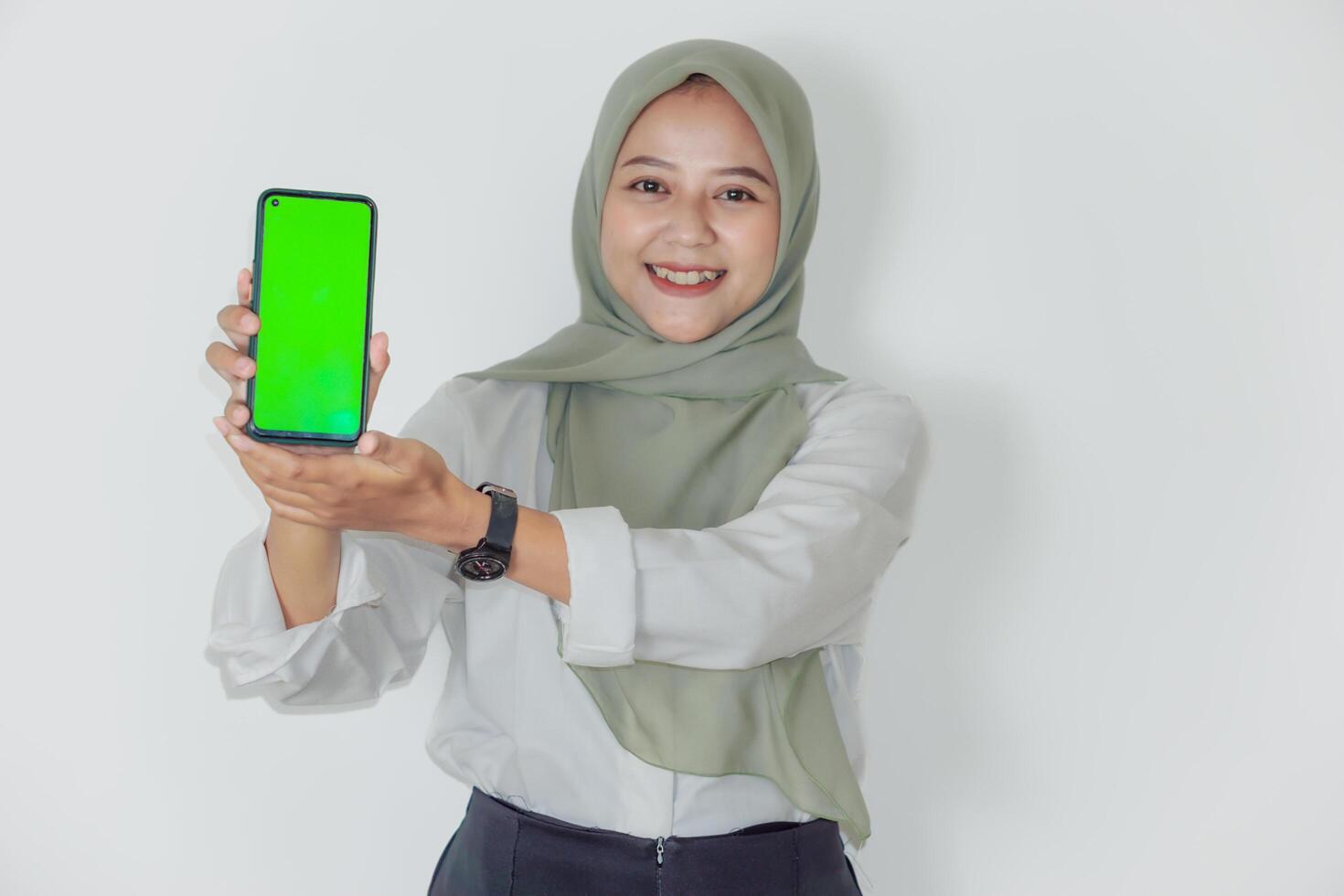 A smiling asian woman in a green hijab holds up a smartphone with a bright green screen. photo