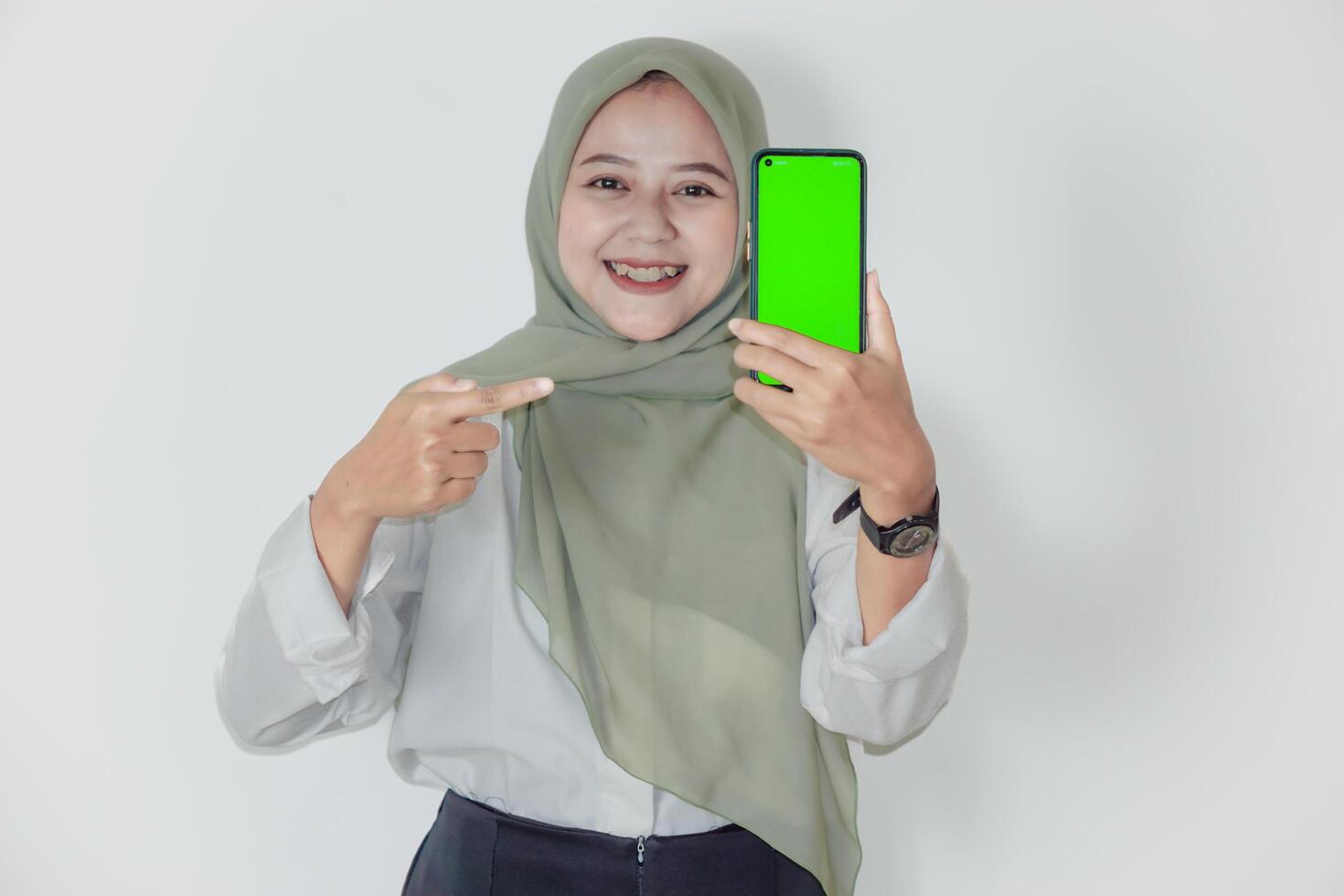 An asian woman in a green hijab smiles while showing her smartphone with a green screen. photo