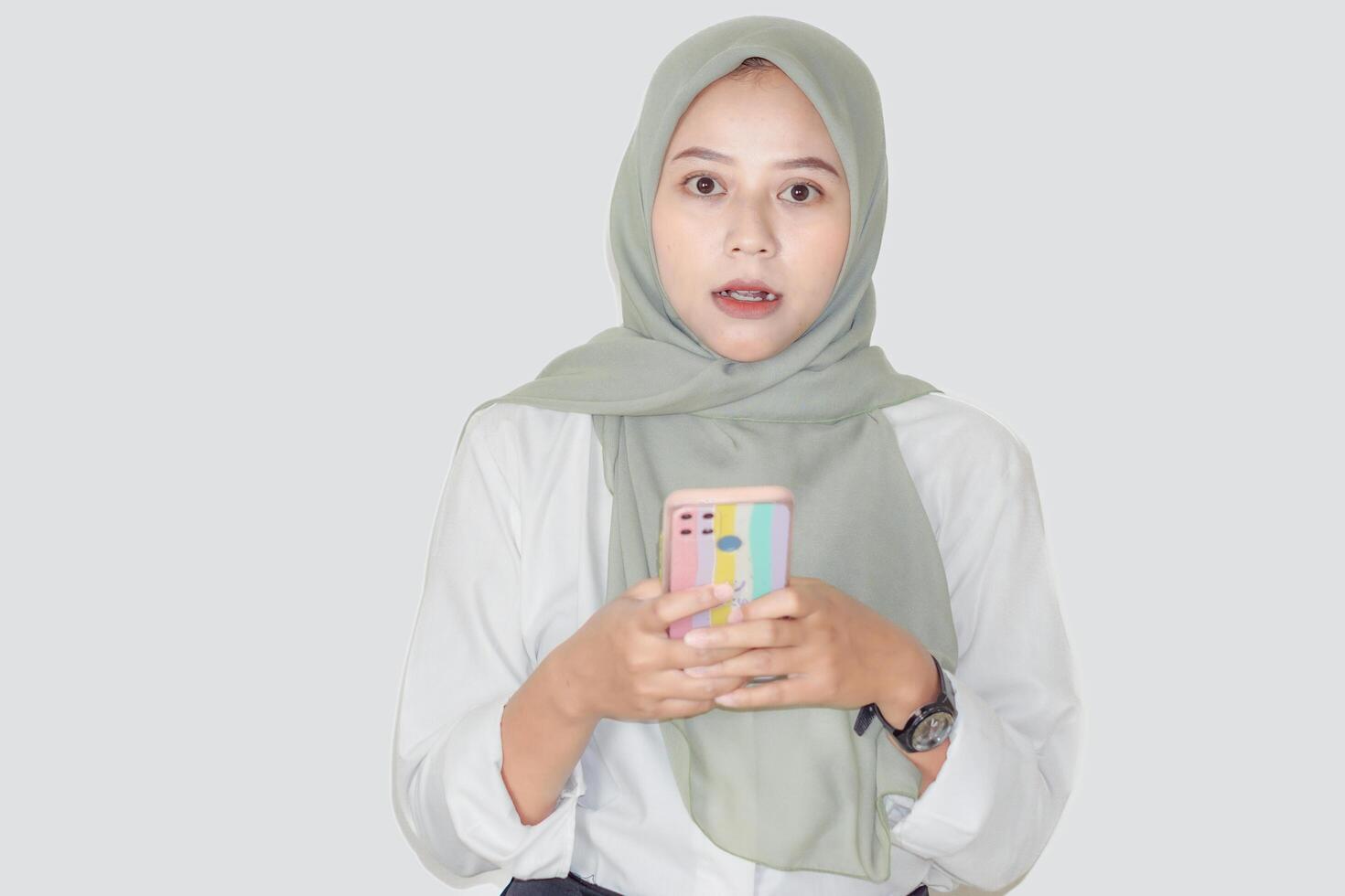 A young asian woman in a green hijab looks surprised while holding her phone, engaged in conversation. photo