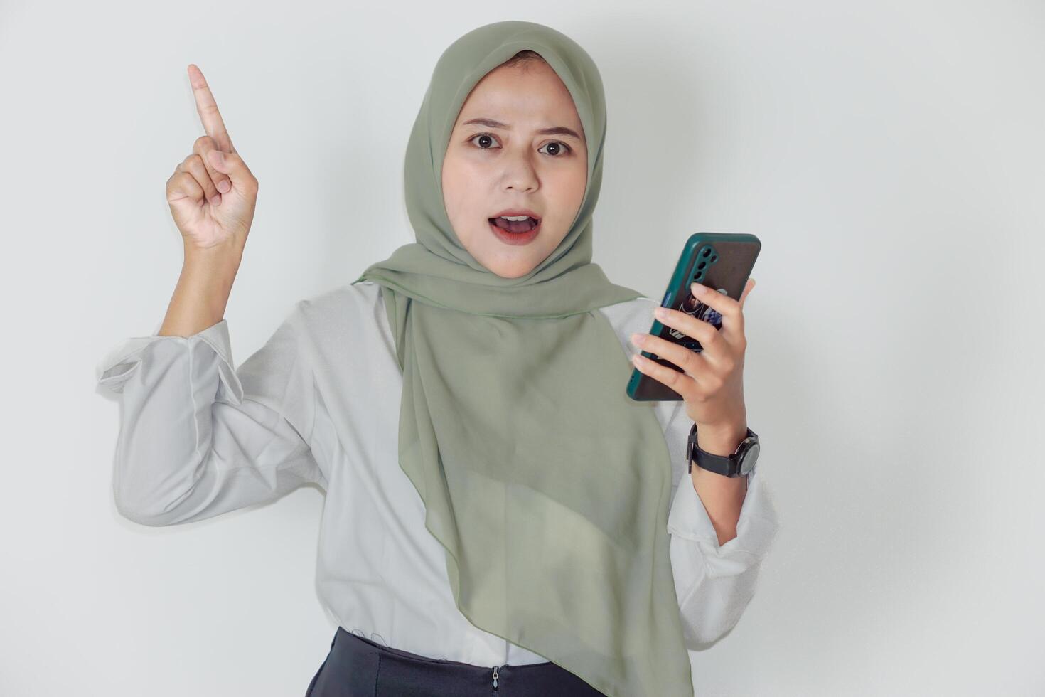 An asian woman in a light green hijab enthusiastically engages while holding a smartphone. photo
